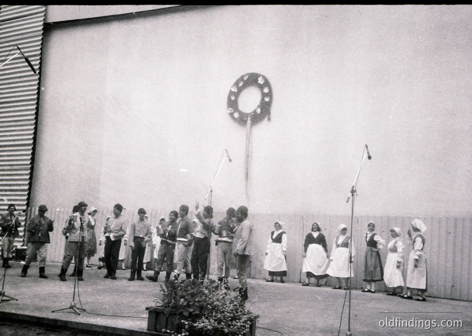 Group singing performance in front of a plain wall, likely a 1960s–70s Eastern Bloc cultural event. Mixed-gender choir in coordinated attire—men in suits, women in aprons—singing near microphones. Decorative wreath on pole suggests a formal or celebratory occasion.