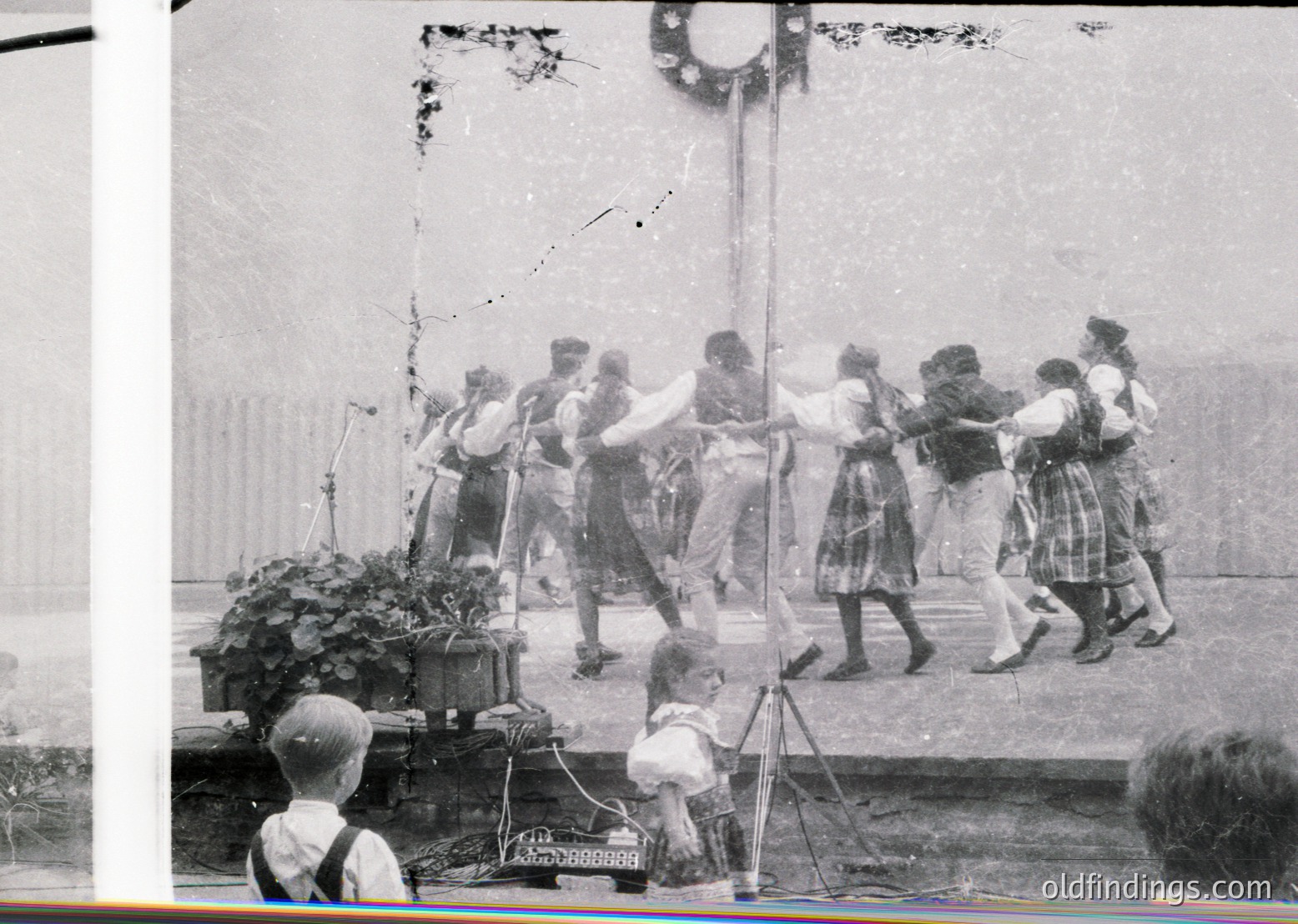 Vintage black-and-white photo of a group dance in an outdoor setting, likely early-to-mid 20th century. Participants in coordinated plaid skirts and blouses form a circle around a decorated pole. A child kneels at the base, observing. Decorative greenery and a sign with Cyrillic script suggest Eastern European cultural event.