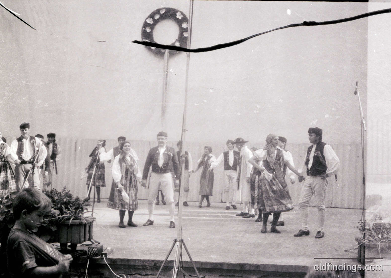 Traditional folk dancers in regional attire performing on an outdoor stage, likely mid-20th century. Men wear vests, hats, and knee-length pants; women in layered skirts and blouses. Stage backdrop features a decorative wheel motif. Crowd and greenery hint at a festive, communal event.