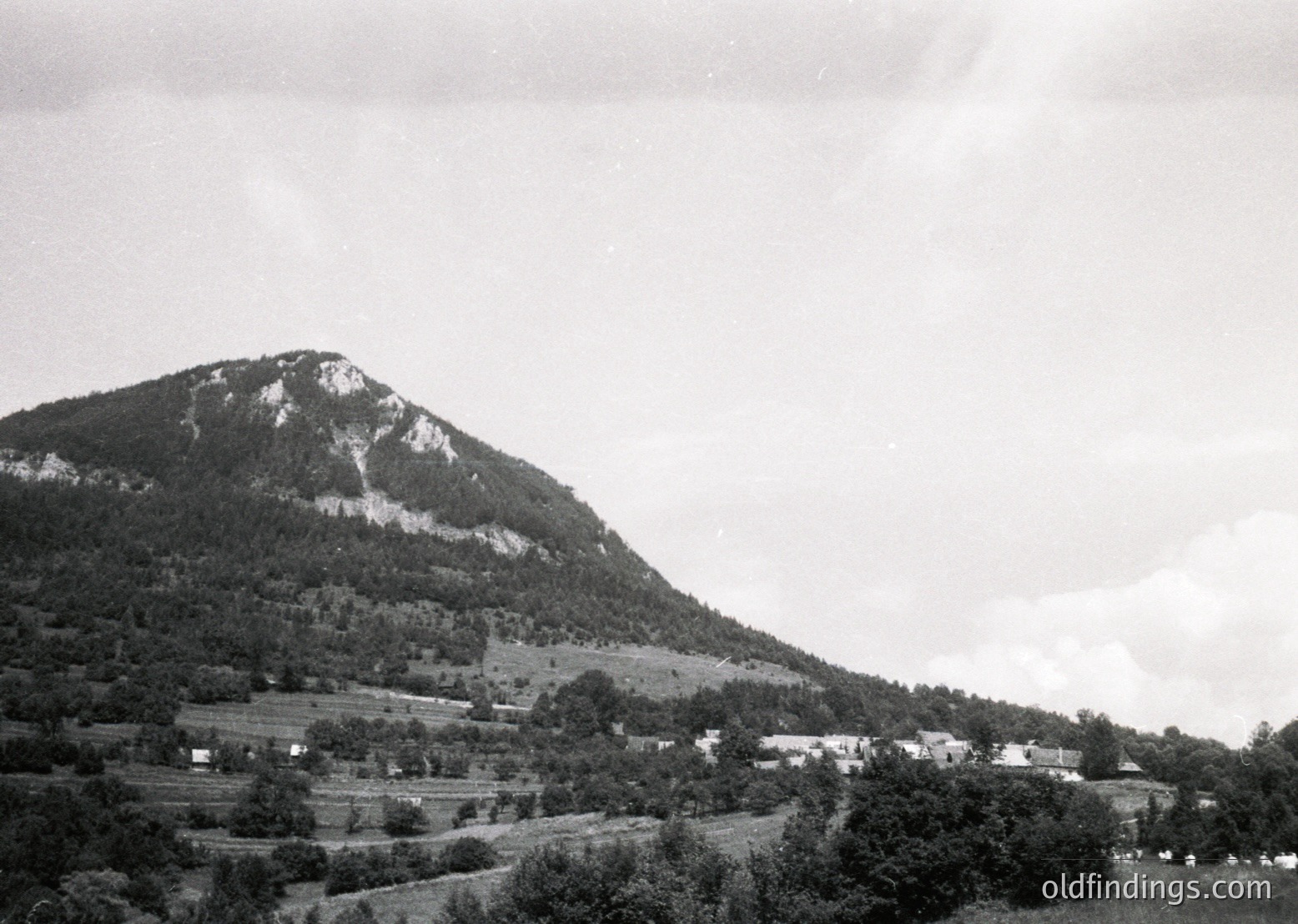 Mid-20th century black-and-white landscape featuring a steep, rocky mountain peak dominating the background. Foreground shows scattered farmhouses and terraced fields, suggesting rural agricultural life. Overcast skies enhance the vintage, timeless feel.