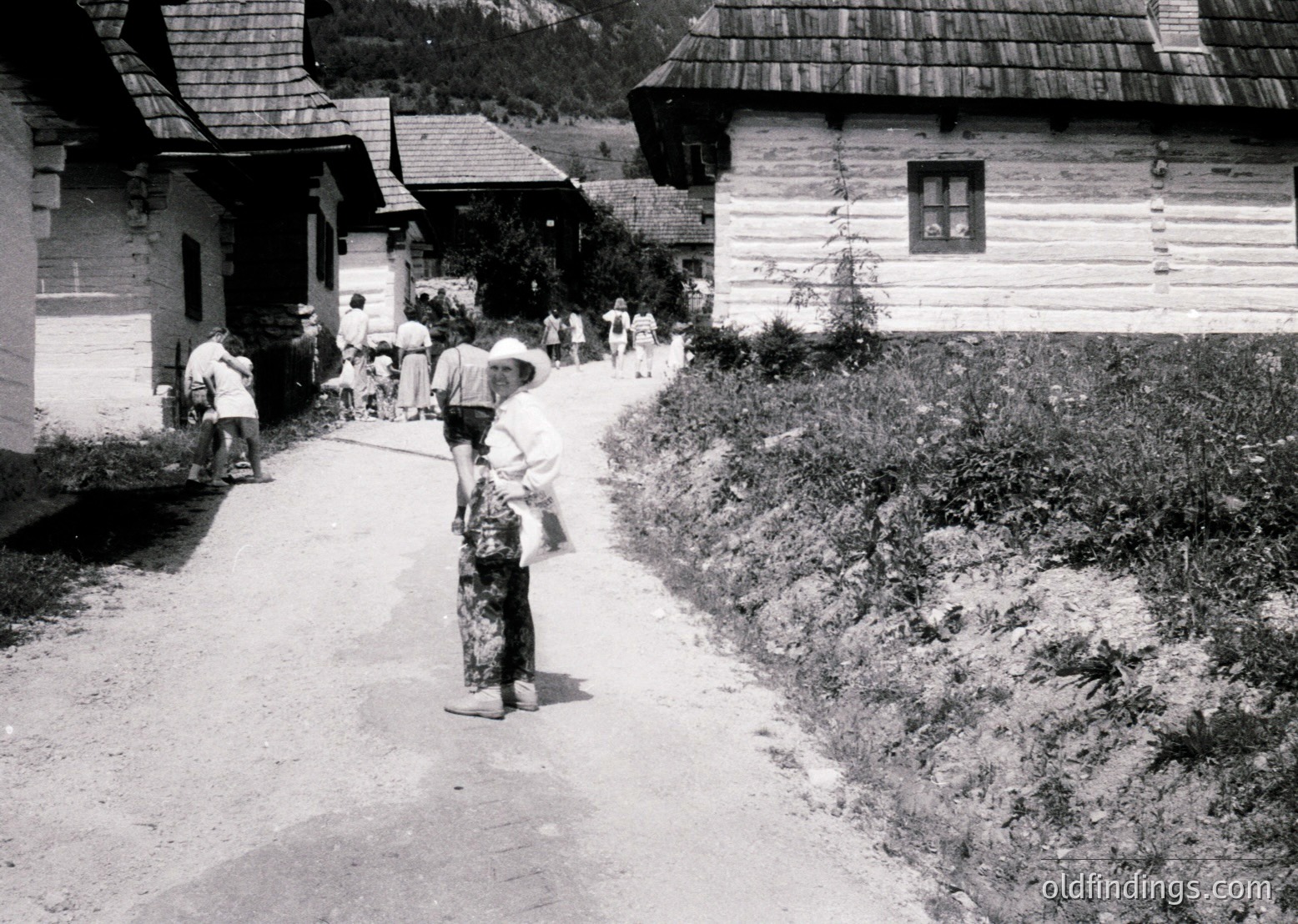 Mid-20th century rural village scene with traditional alpine-style wooden houses. A woman in patterned dress and headscarf leads a child by hand along a dirt path. Group of people, likely locals, gather near the buildings.