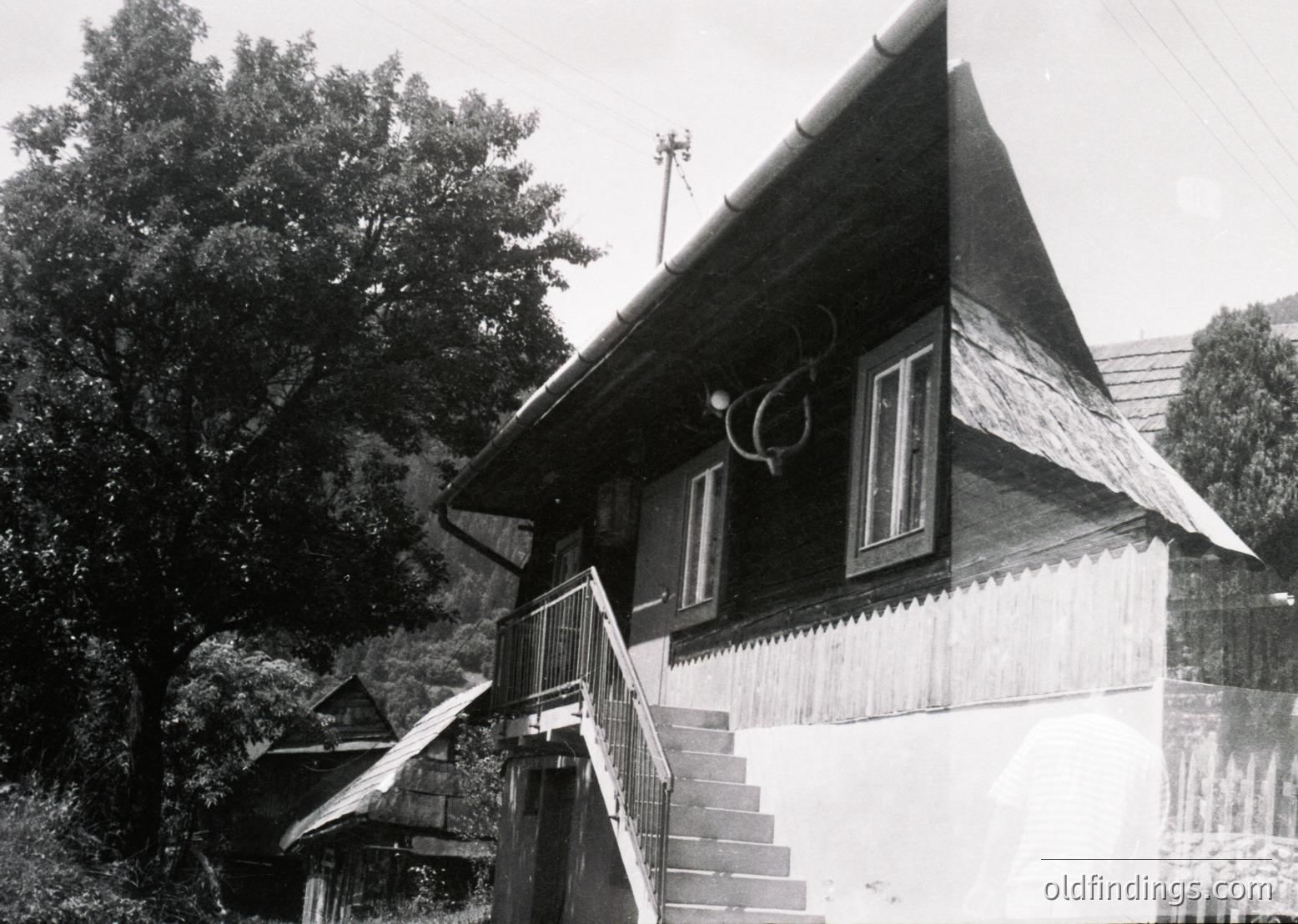 Mid-20th century wooden house with steep gable roof and decorative trim, set in a forested area. Exterior stairs lead to an elevated entrance, framed by a small balcony. Rustic architectural details suggest rural European design, likely –.
