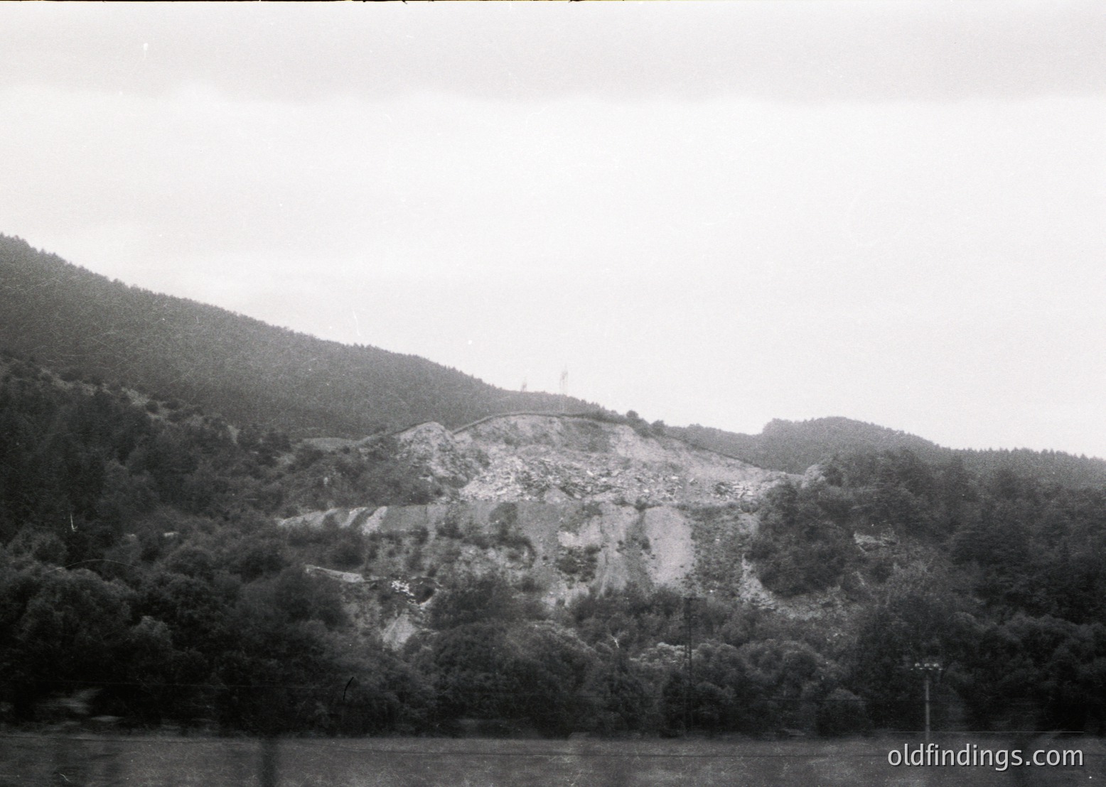 Black-and-white landscape featuring a rocky, terraced hillside with sparse vegetation, likely post-industrial or agricultural. Water body in foreground suggests a lake or river. Dense forest covers lower slopes. Mid-20th century style suggests historical or archival context.