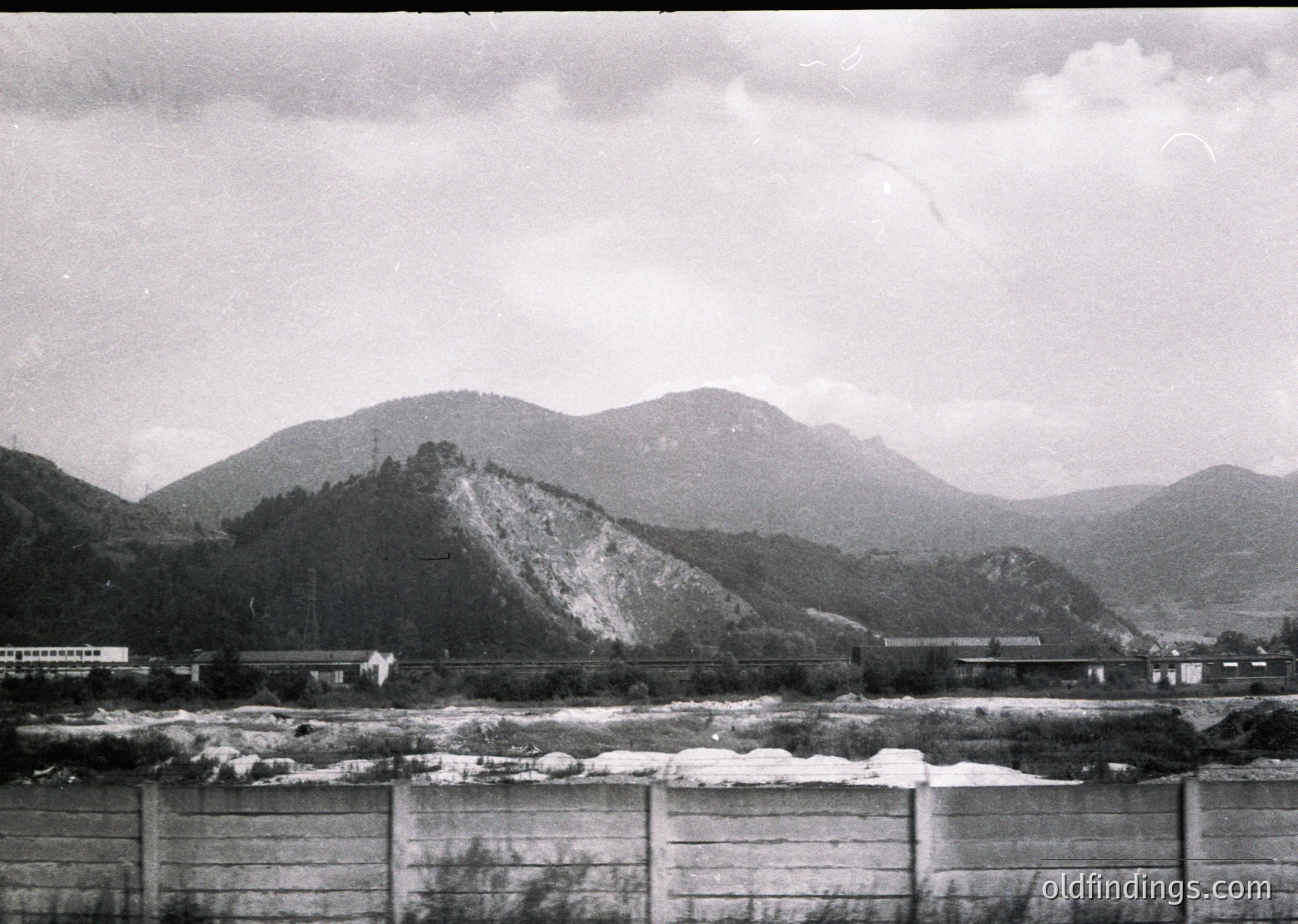 Mid-20th century black-and-white landscape featuring a dam with snow-capped rocks below and forested mountains in background. Industrial buildings and power lines hint at hydroelectric development.
