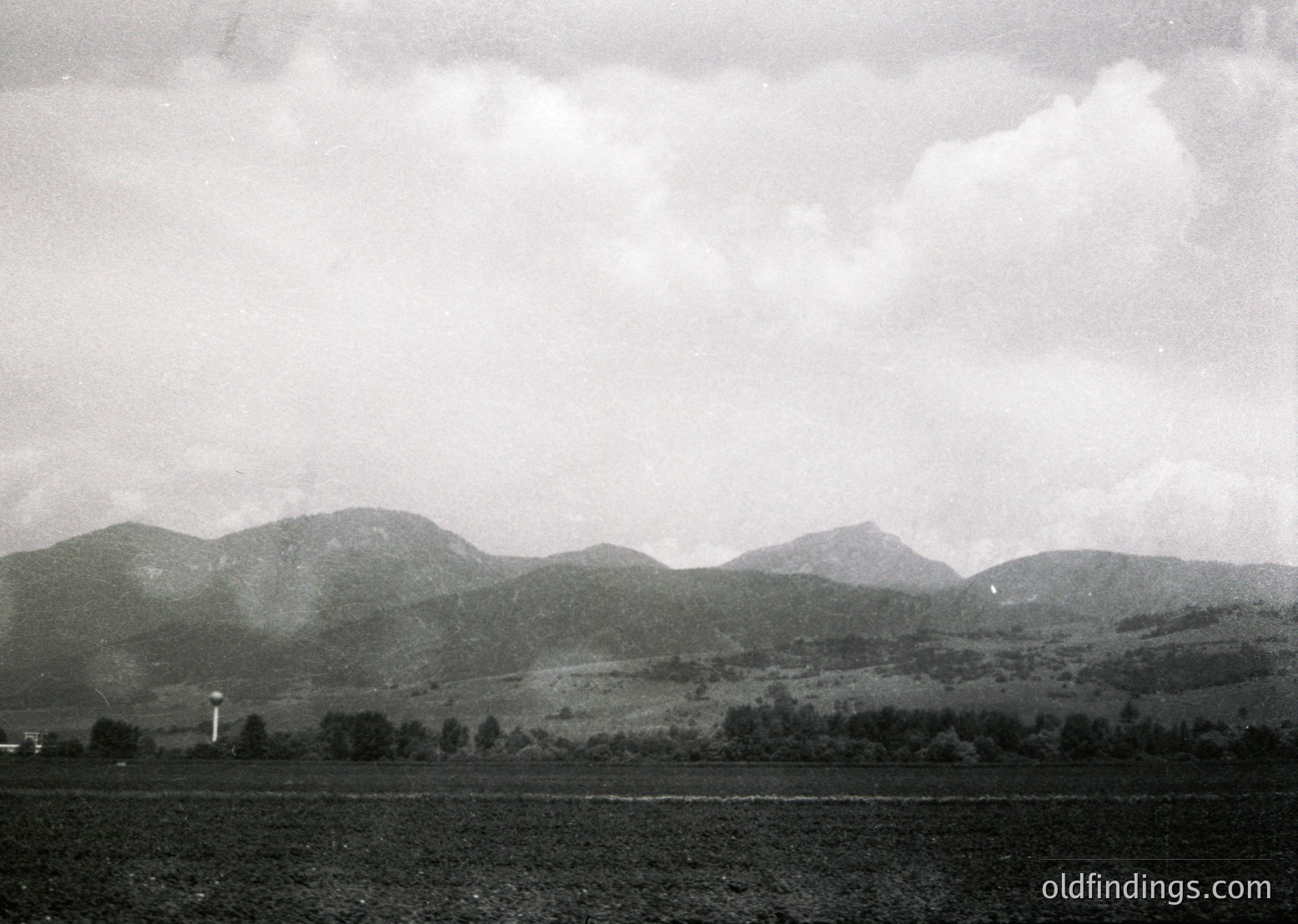 Vintage black-and-white landscape of rolling hills and farmland under overcast skies. Distant radio tower and sparse trees mark horizon. Mid-20th century rural European countryside, likely Eastern Bloc era.