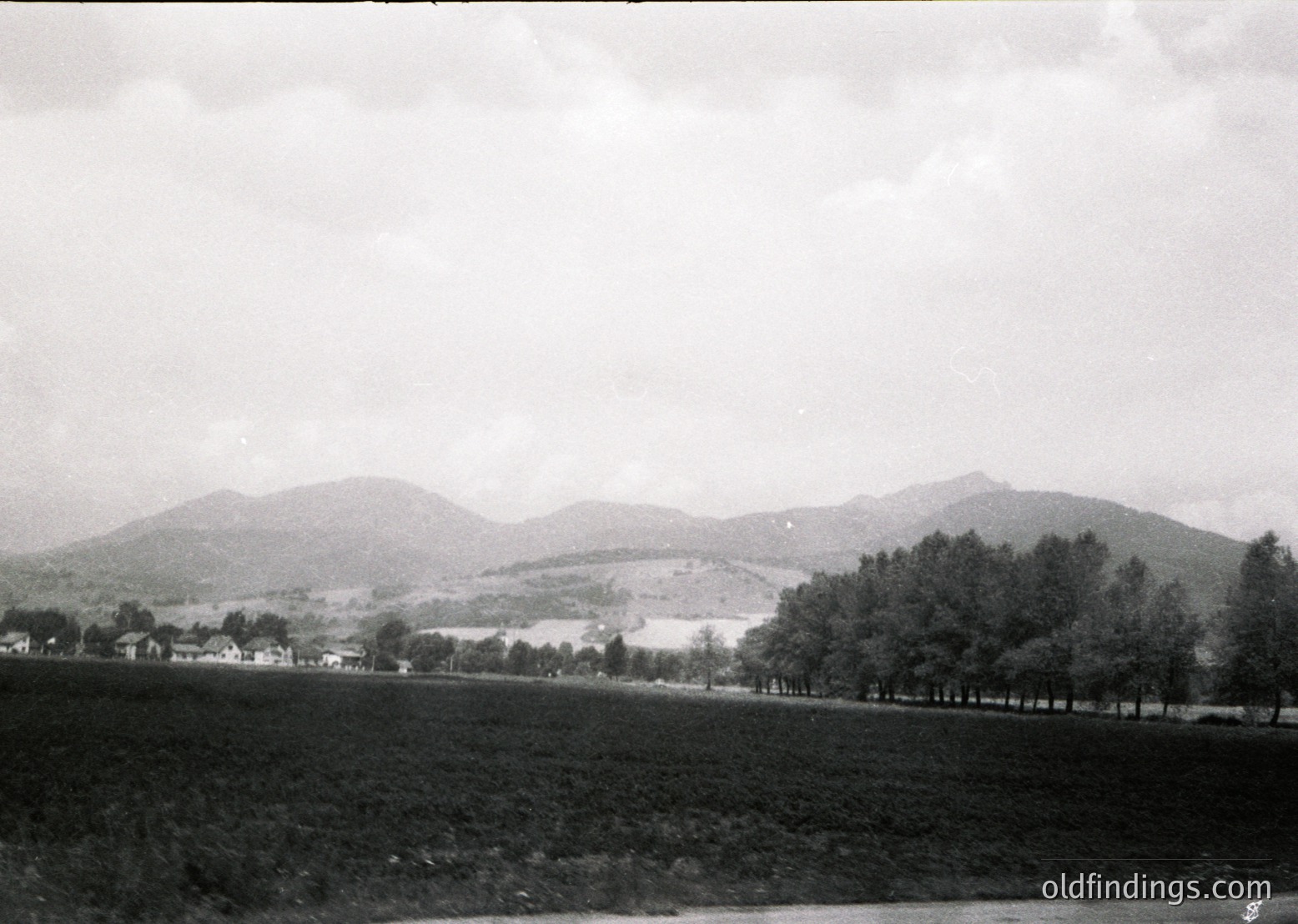 Black-and-white rural landscape featuring rolling hills, dense tree line, and scattered farmhouses. Mid-20th century agricultural setting with plowed fields and distant settlement.