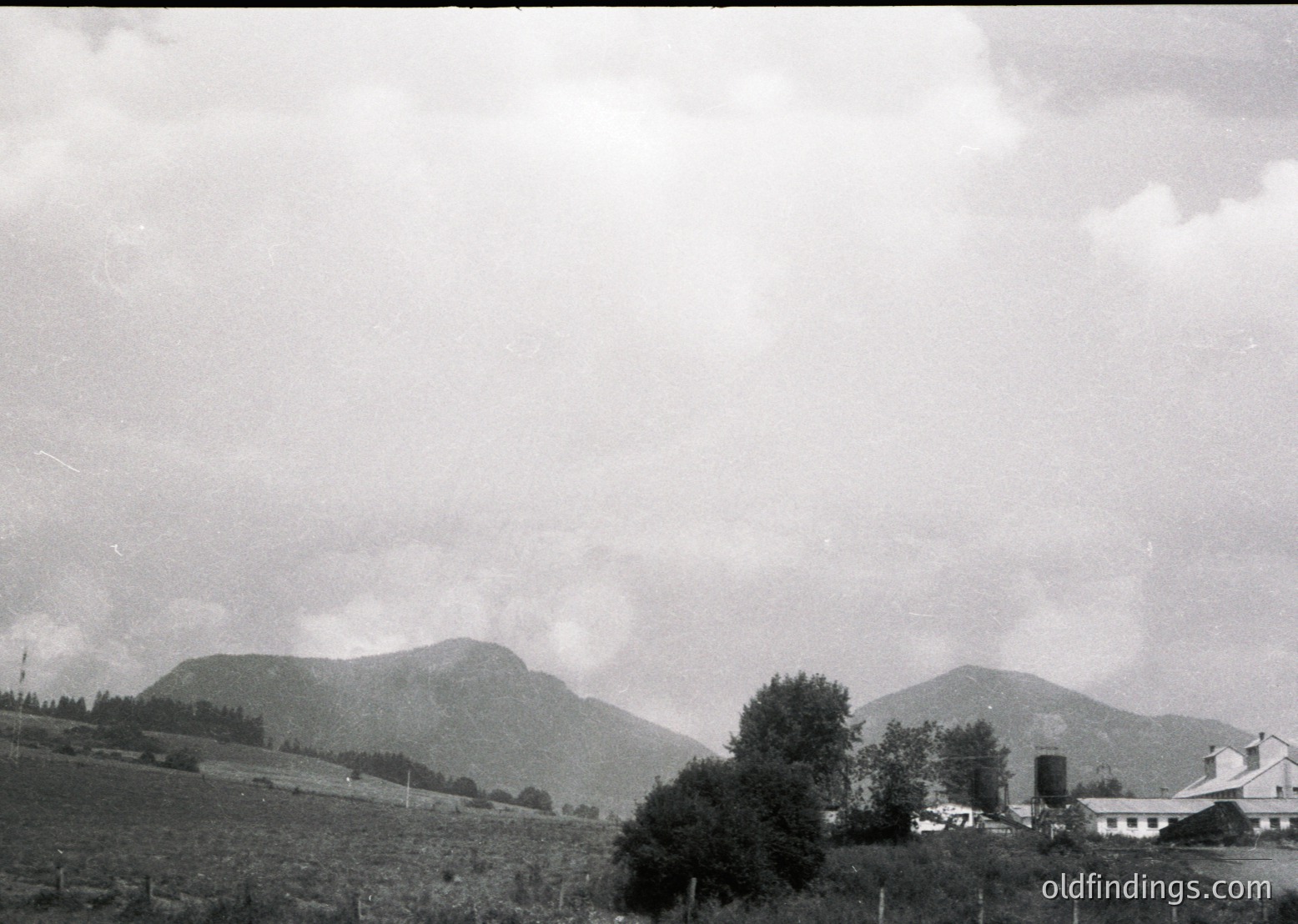 Vintage black-and-white landscape featuring rolling hills, dense forest clusters, and a lone white farmhouse with a cylindrical water tower. Mid-20th century rural European countryside, likely .