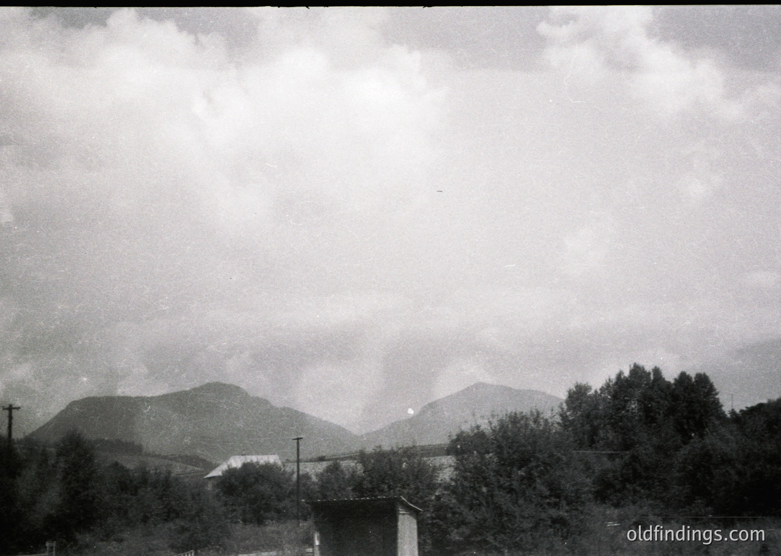 Mid-20th century black-and-white landscape featuring rolling hills and dense forest under overcast skies. A lone utility pole and small concrete structure (likely a guardhouse or shelter) frame the lower right. Dramatic cloud formations suggest impending rain.