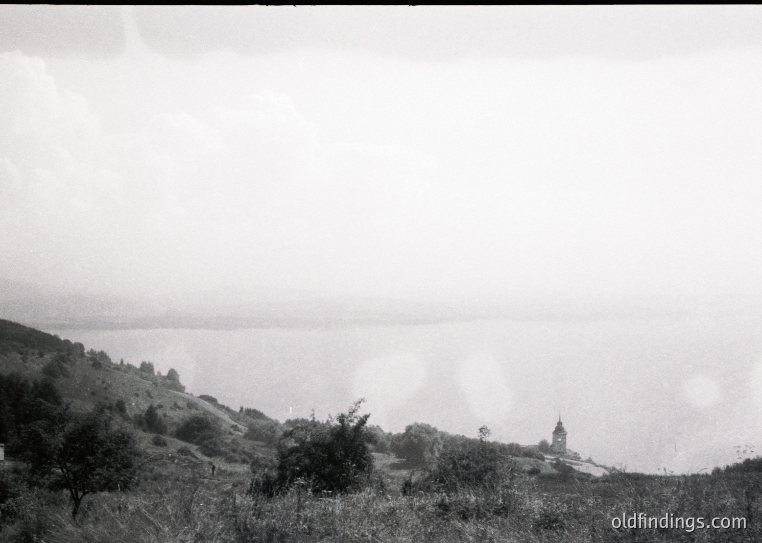 Vintage black-and-white coastal landscape with misty horizon and distant church spire. Rolling hills frame a serene seaside view, likely Eastern European. Early-to-mid 20th century composition.