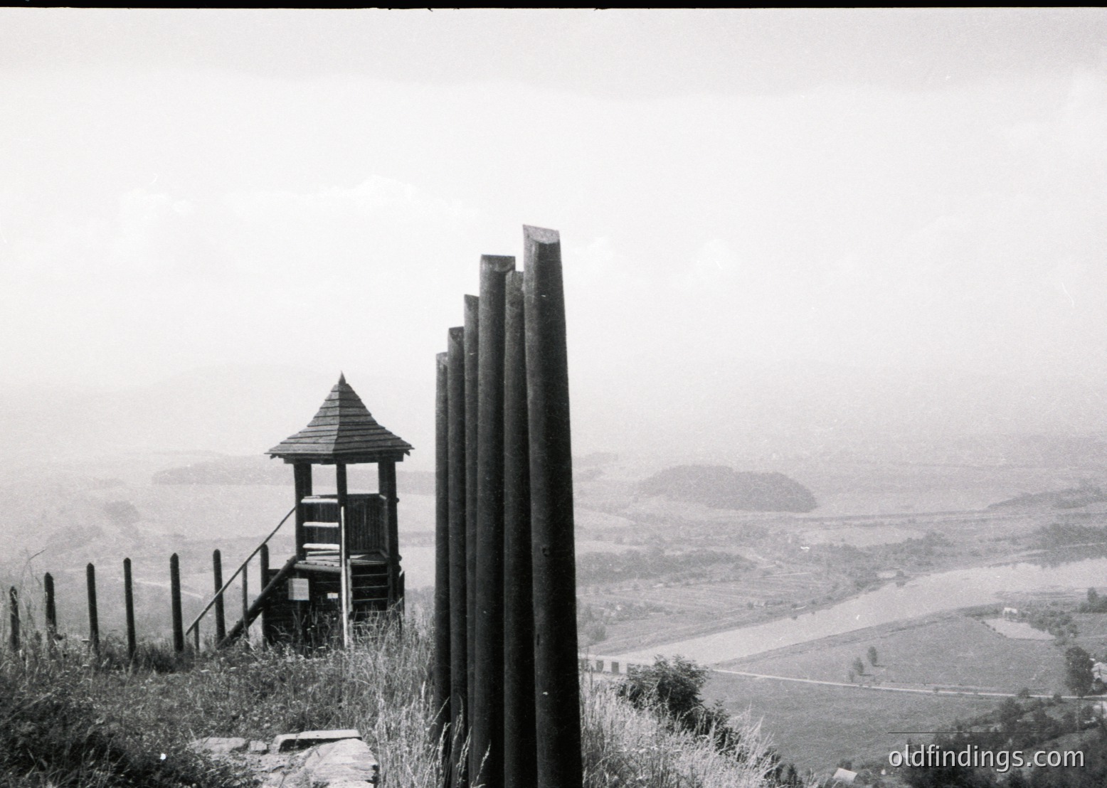 Rustic wooden observation tower perched on a hillside overlooking a winding river valley, framed by vertical wooden posts. Mid-20th century rural architecture with simple, functional design. Misty landscape suggests early morning or overcast conditions.