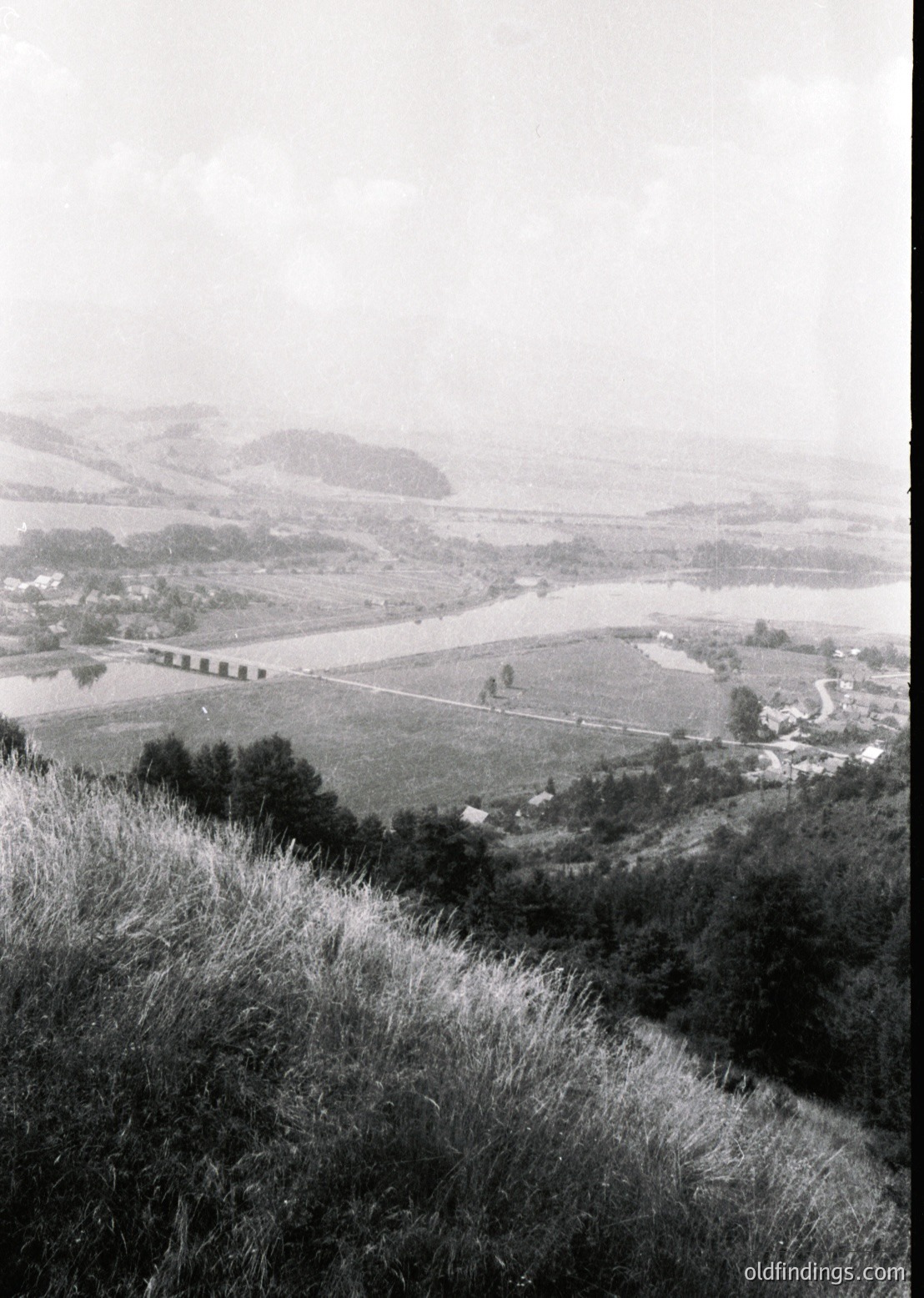 Mid-20th century black-and-white landscape featuring a winding river valley with agricultural fields, dense forest patches, and a distant road bridge. Mist obscures higher elevations, enhancing atmospheric depth. Rural setting likely in a temperate climate zone.