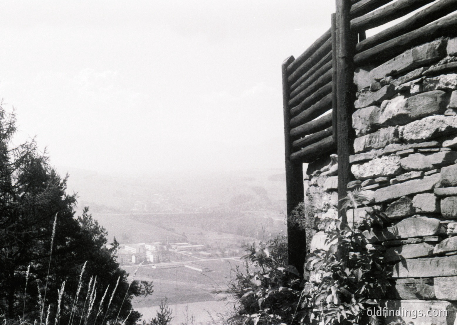 Black-and-white rural scene featuring a stone wall with wooden fence atop a misty valley. Distant farmhouses and fields stretch below, framed by dense forest. Likely European alpine or mountainous region, mid-20th century.