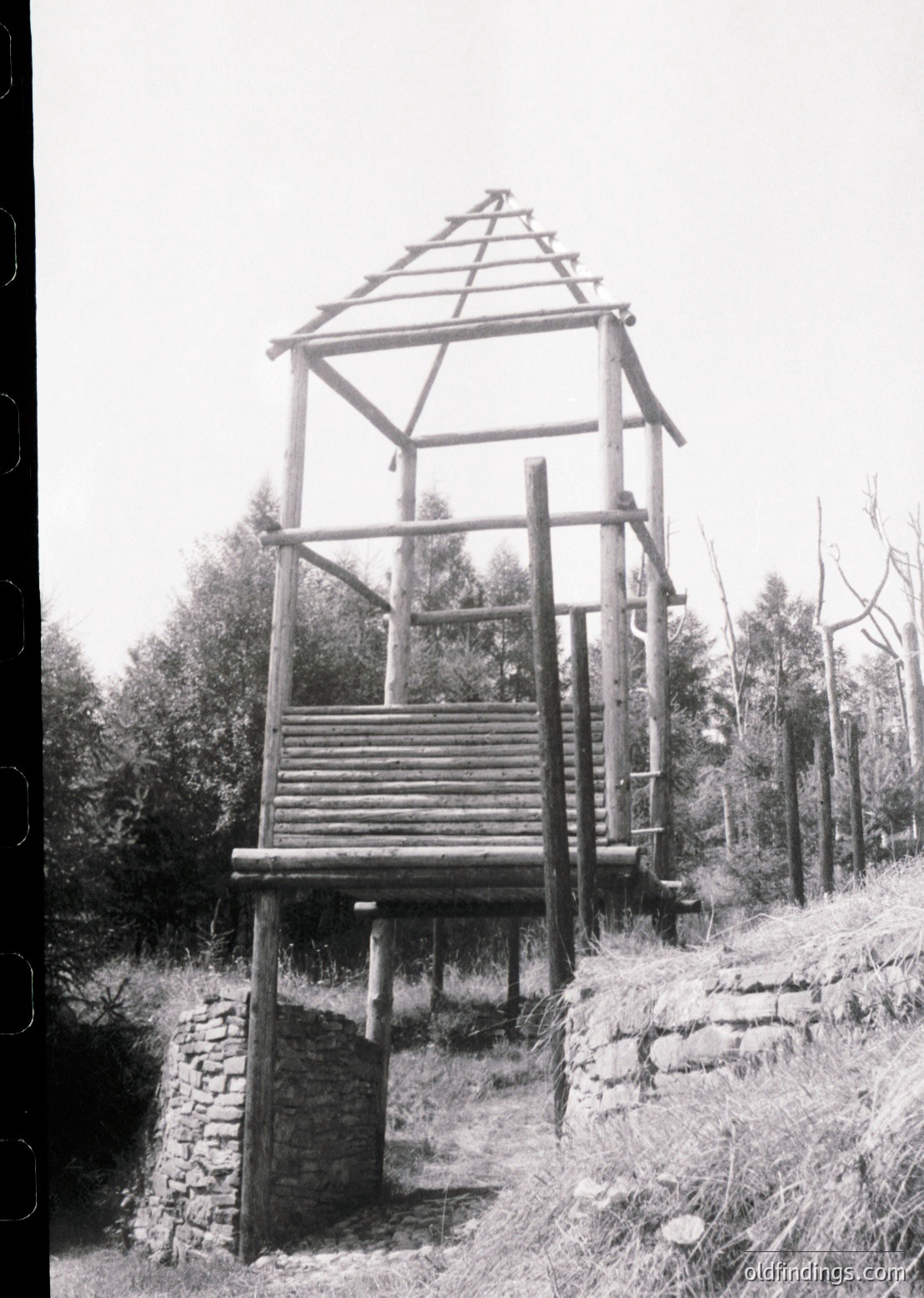 Traditional wooden water tower with steep gabled roof, elevated on stone foundation. Rustic design suggests rural European heritage, likely 19th–early 20th century. Surrounded by dense forest and stone pathway.