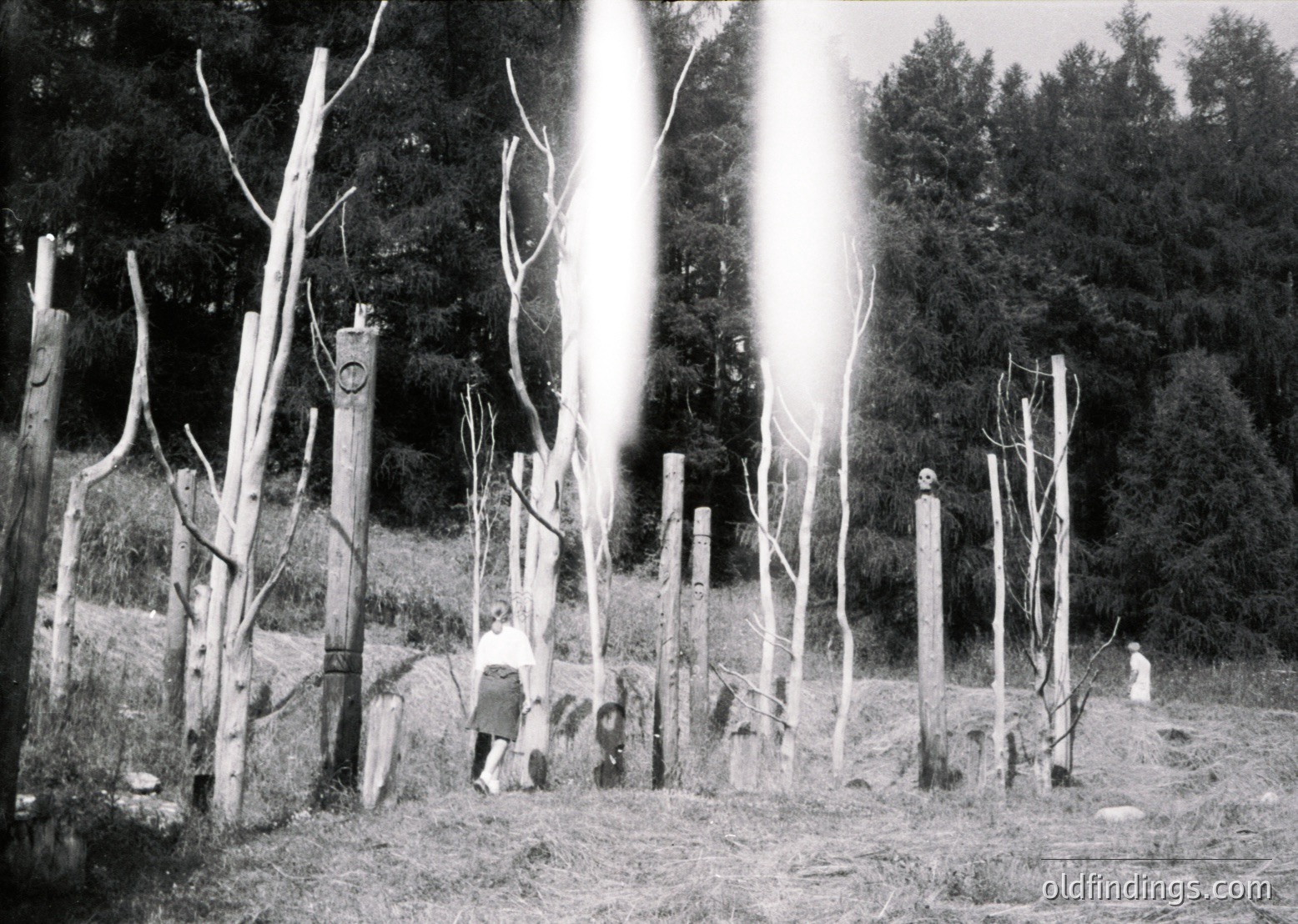 Black-and-white rural scene featuring a wooden fence with vertical posts and horizontal rails, likely constructed for livestock containment. Two individuals—one adult and one child—walk near the fence, dressed in long-sleeve shirts and dark pants. Sunlight filters through bare trees, creating dramatic backlighting. The setting appears to be a farm or agricultural area, possibly mid-20th century.