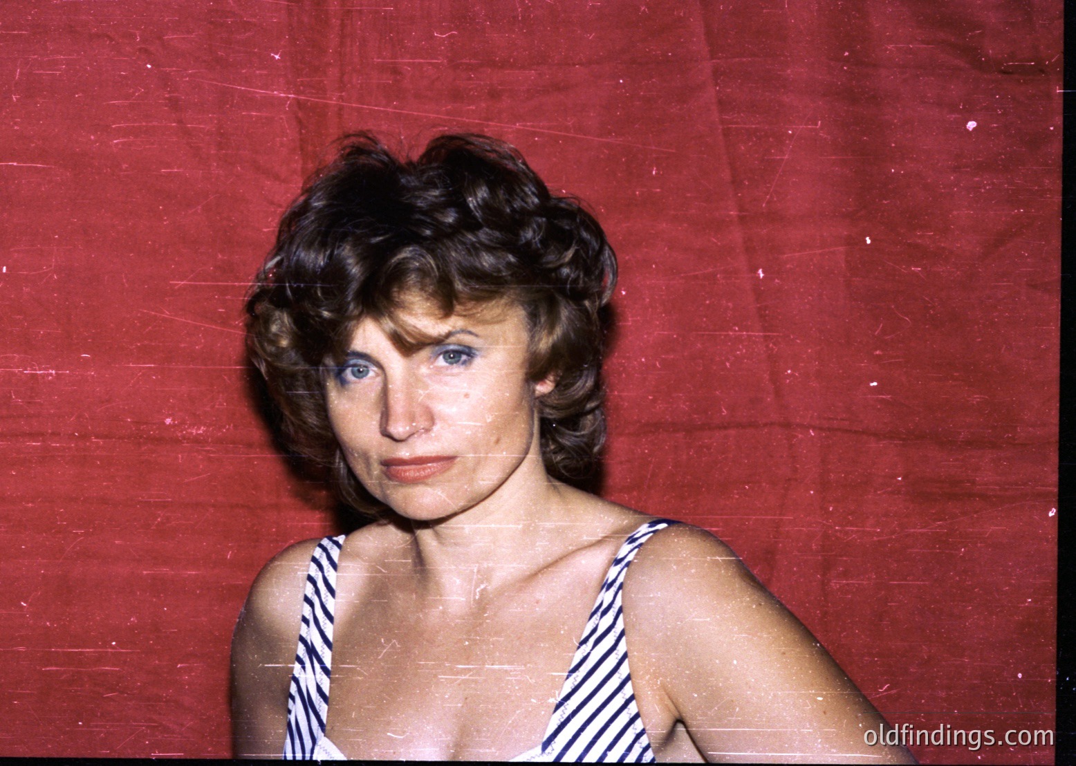 Portrait of a woman in a striped swimsuit against a red backdrop, likely a 1960s–1970s beach or resort setting. Her short, voluminous hairstyle and bold makeup reflect mid-century fashion trends.