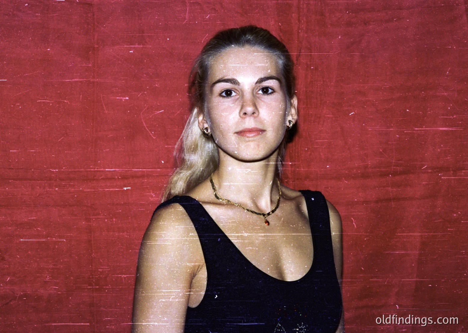 Portrait of a woman with short, straight blonde hair in a sleek bob, wearing a black sleeveless dress and gold earrings. The vintage sepia-toned background suggests an indoor setting, possibly a studio or event space. Style reflects 1970s–1980s fashion trends.