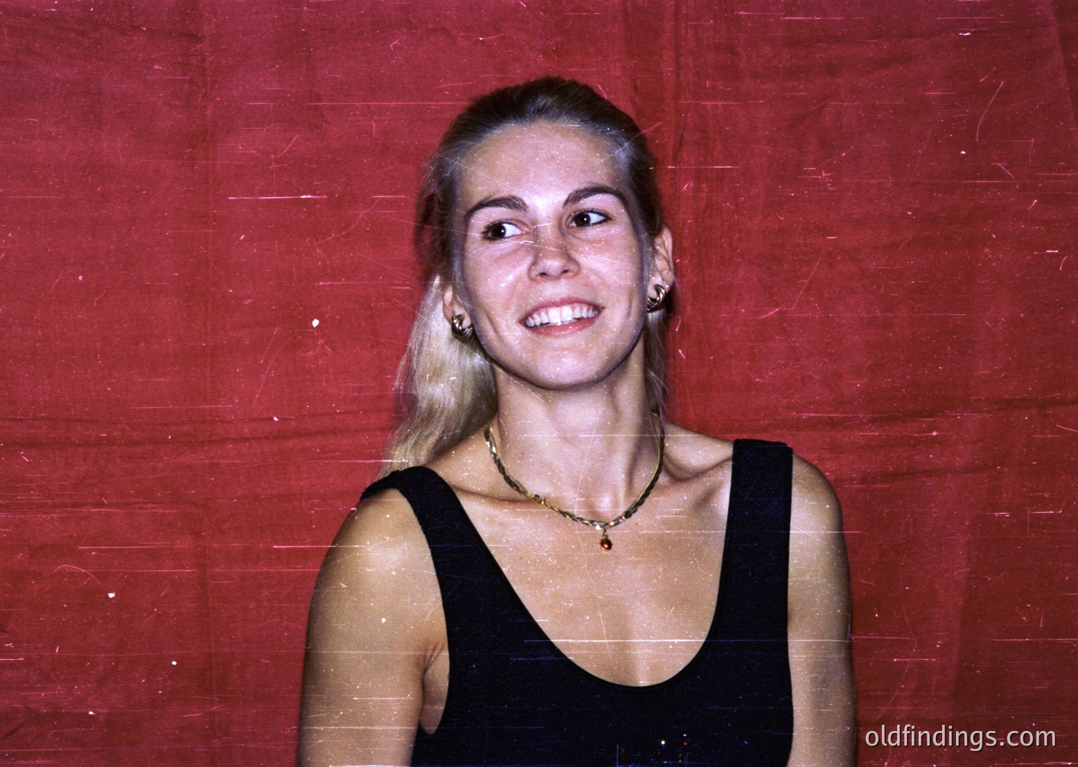 Vintage portrait of a woman in a sleeveless black dress, set against a faded red backdrop. Her blonde hair is shoulder-length, parted in the middle, with a subtle side part. She wears minimal jewelry: a thin necklace and small earrings. Likely late 20th-century fashion, possibly or .