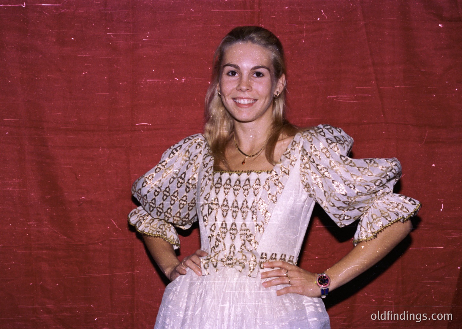 Vintage portrait of a woman in a traditional embroidered blouse with puffed sleeves, paired with a flowing skirt. The patterned fabric features geometric and floral motifs, suggesting Eastern European heritage. Warm lighting and a red backdrop enhance the retro aesthetic, likely from the 1970s–1980s.