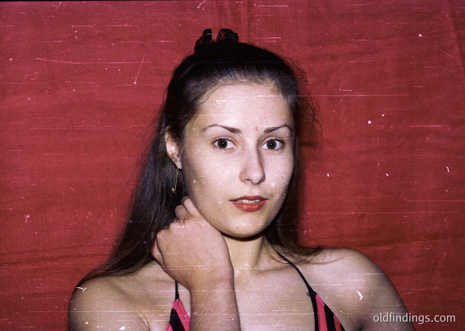 Portrait of a woman in a vintage-style swimsuit against a faded red backdrop, likely from the 1970s. Her hair is pulled back with a black headband, and she wears minimal jewelry. The image evokes mid-century fashion and candid photography.