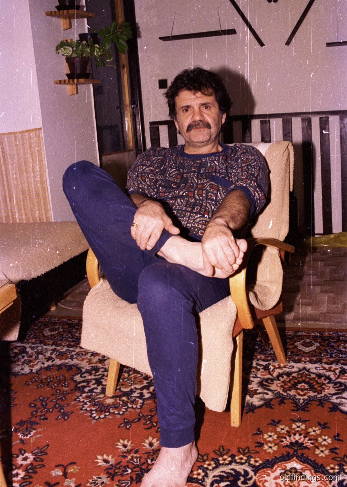 Mid-century interior portrait of a man in a patterned sweater and wide-legged trousers, seated in a wooden armchair on a floral rug. Geometric wall patterns and potted plants suggest a 1960s–1970s European home. Warm lighting enhances vintage aesthetic.
