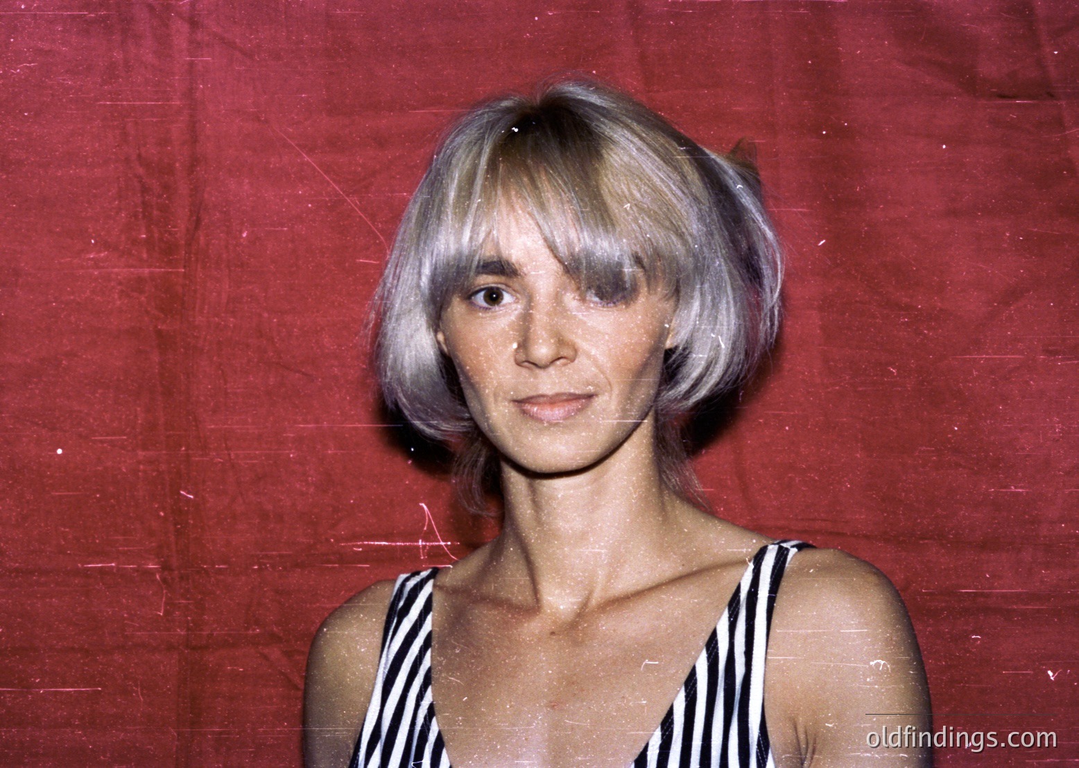 Portrait of a woman with short, layered blonde hair in a 1970s-inspired bob, set against a maroon backdrop. She wears a striped sleeveless top, exuding a retro aesthetic. Likely a candid or studio shot from the 1970s–1980s, evoking mid-century fashion trends.