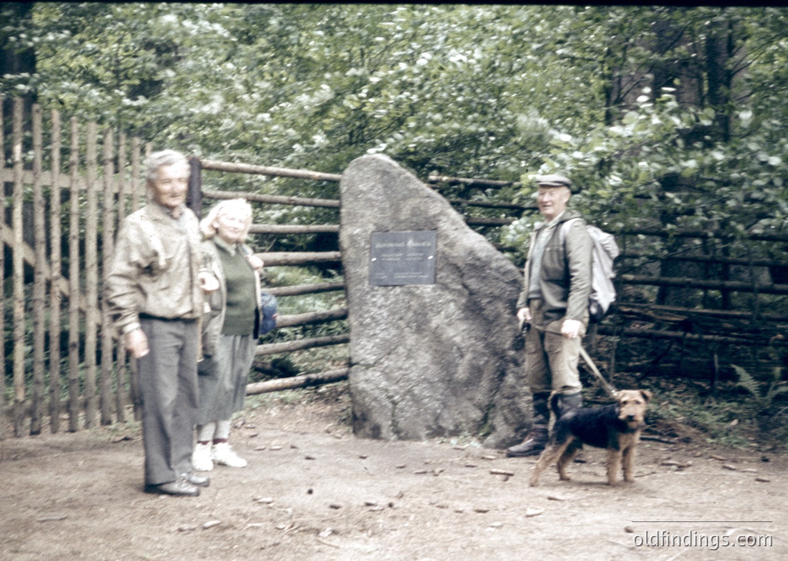 Mid-20th century outdoor scene with three adults and a dog near a wooden fence and large boulder plaque. The man on the right wears hiking gear, including a backpack and hat, suggesting a nature excursion. The sepia-toned photo captures a rustic, forested setting, likely from the 1950s–1960s.