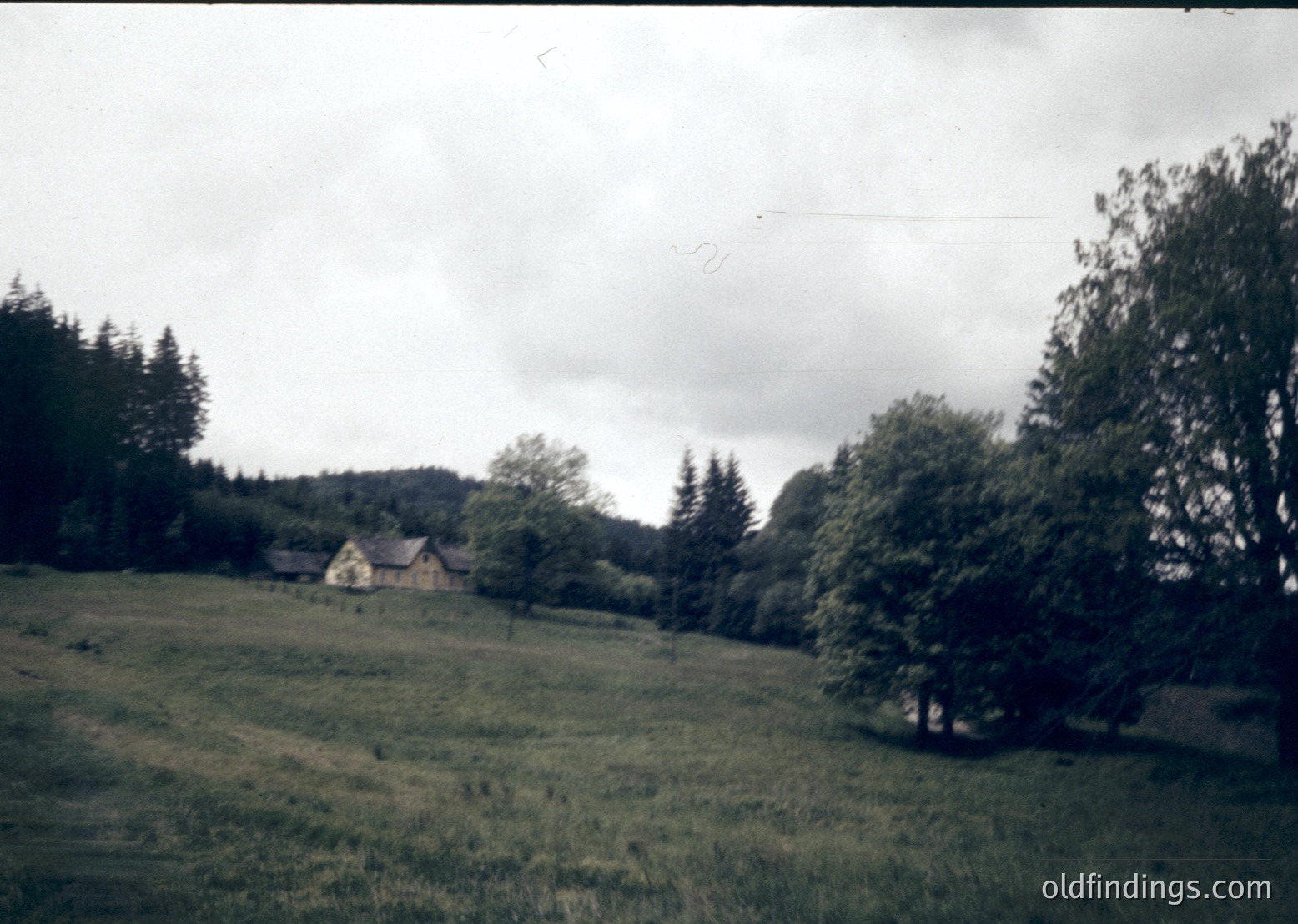 Vintage sepia-toned rural landscape featuring a single-story stone farmhouse with a pitched roof, nestled in a lush, rolling meadow. Dense forest and rolling hills frame the horizon under an overcast sky. Likely European countryside, mid-20th century.