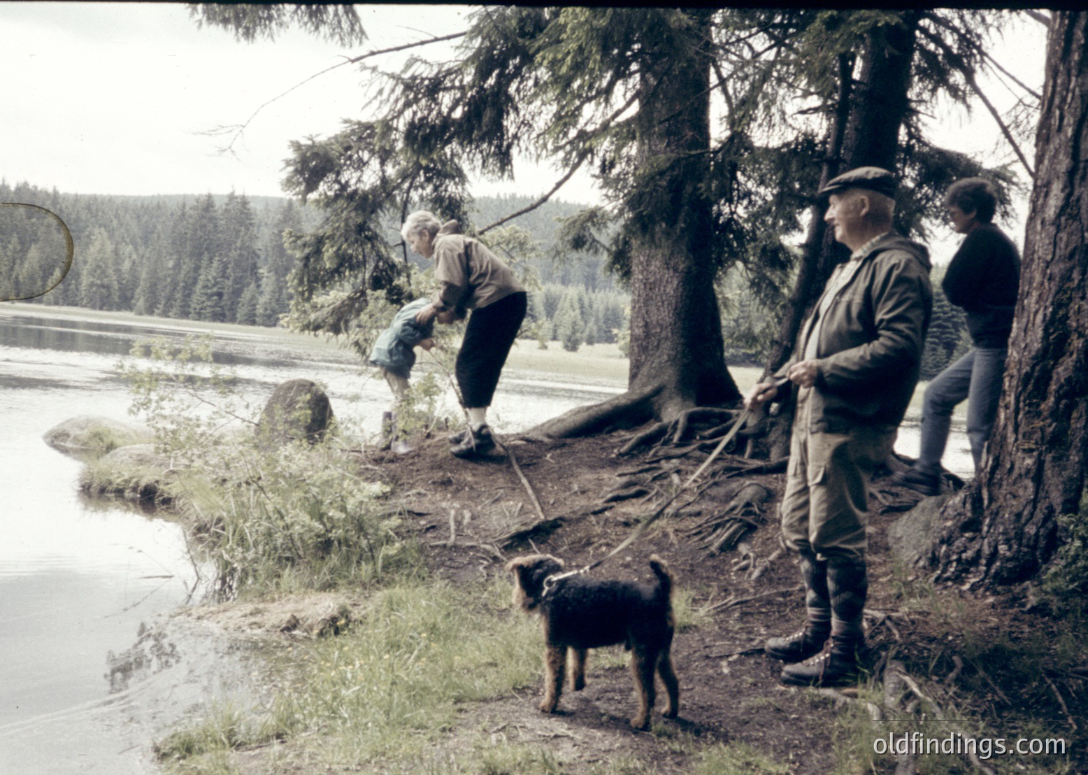 Vintage black-and-white photo of three men and a dog near a forested lakeside, likely mid-20th century. Men in practical outdoor attire—one bending to assist a child, another holding a stick, and a third standing with a dog on a leash. Dense pine trees and calm water reflect rural, alpine lifestyle.