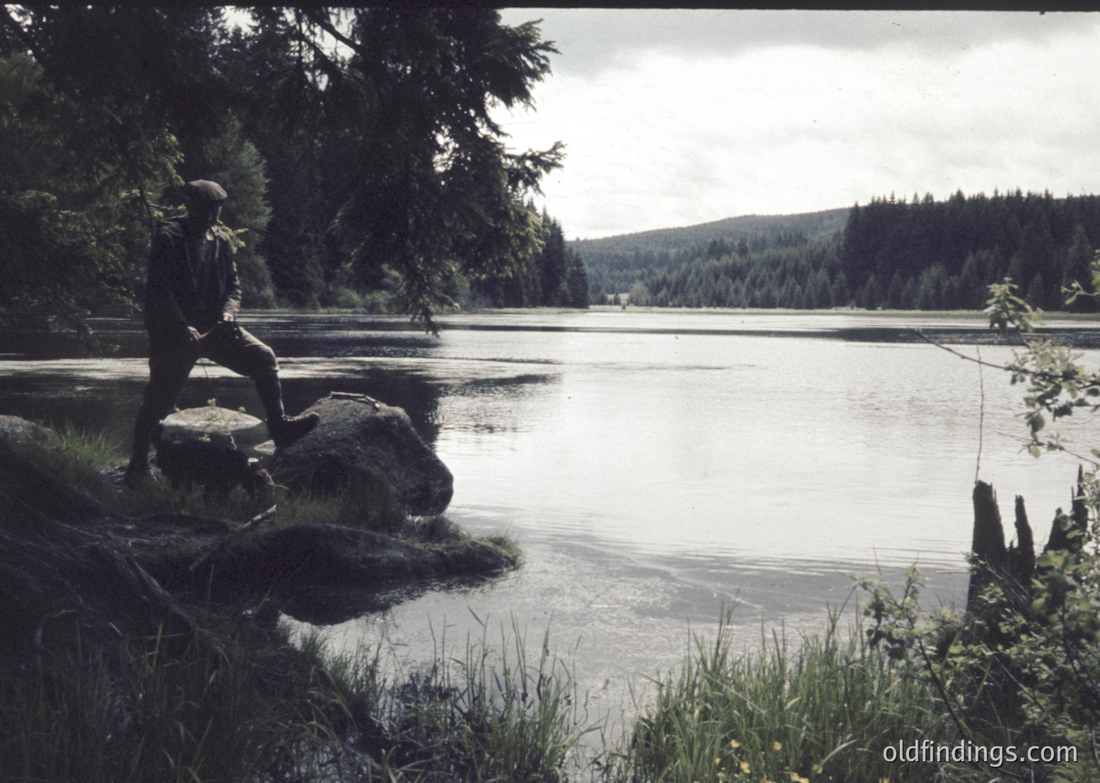 A lone figure sits on a rock by a serene lake, surrounded by dense forest. Mid-20th century outdoor photography captures the rugged natural setting with calm waters reflecting forested hills.