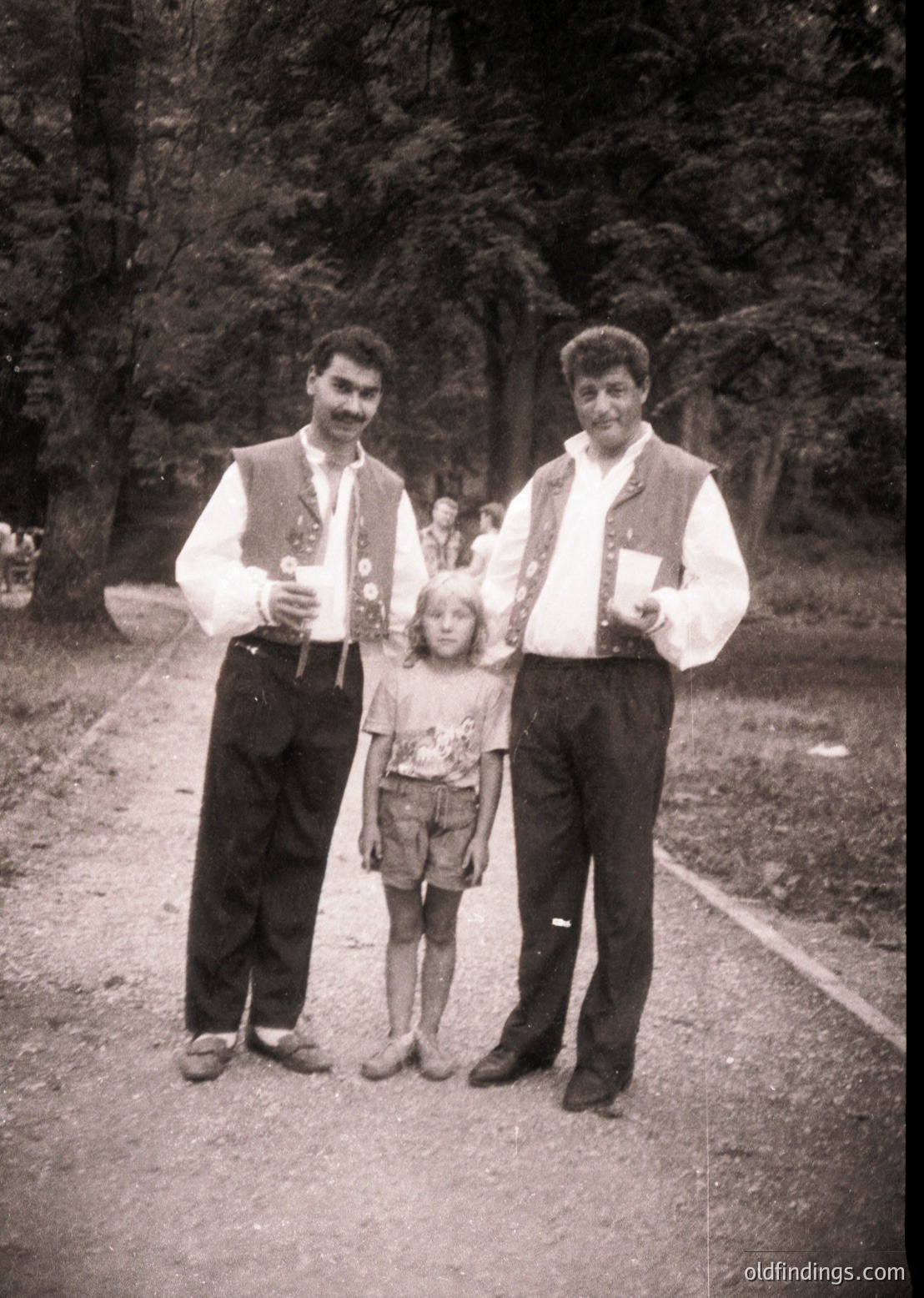 Family portrait in traditional folk attire, mid-20th century. Two men in embroidered vests and knee-high boots flank a young girl in a floral dress, holding small wooden figurines. Wooden path and forested background suggest rural setting.