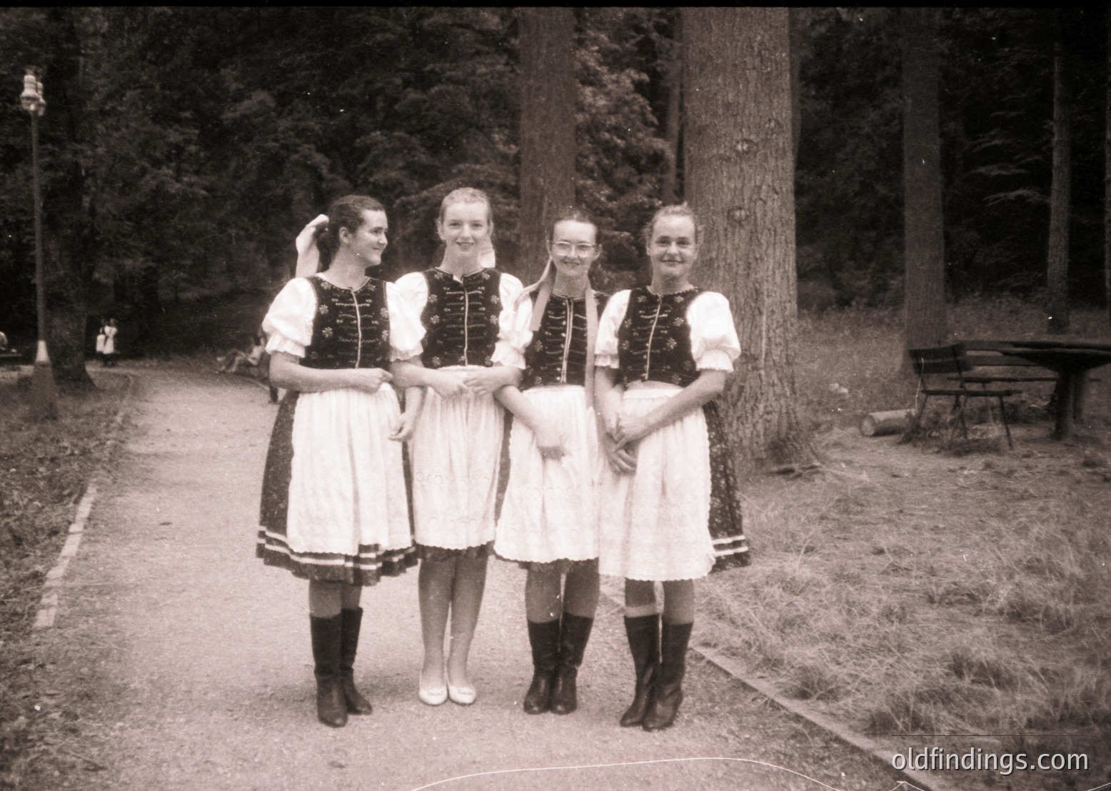 Four women in traditional Alpine folkwear pose on a gravel path, surrounded by dense forest. White blouses with dark embroidered vests, black knee-high boots, and white aprons with lace trim. Mid-20th century (1950s–1960s) European rural setting.