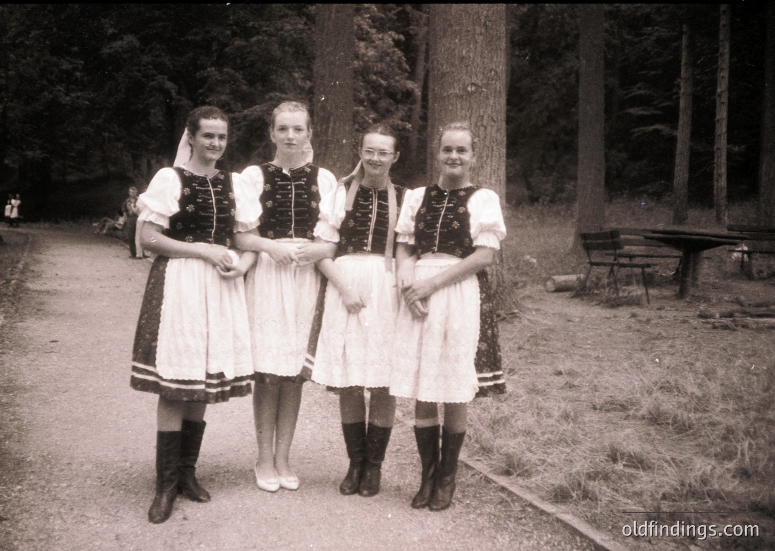 Four women in traditional Alpine folkwear pose outdoors, likely mid-20th century. White blouses with black embroidered vests, knee-length skirts, and knee-high boots. Forest path and wooden bench in background.