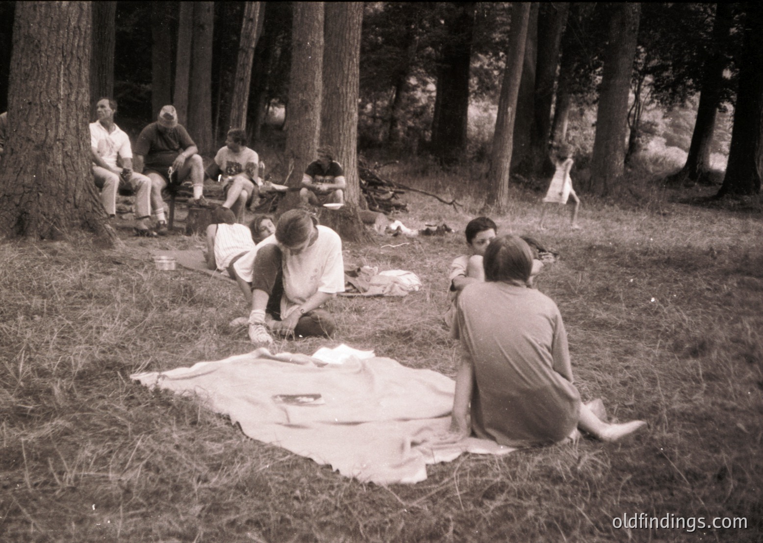 Vintage black-and-white group picnic in a dense forest clearing, mid-20th century. Seven individuals seated/lying on a blanket, surrounded by tall trees and scattered foliage. One person kneels, another reclines on the blanket. Casual summer attire suggests leisure activity.