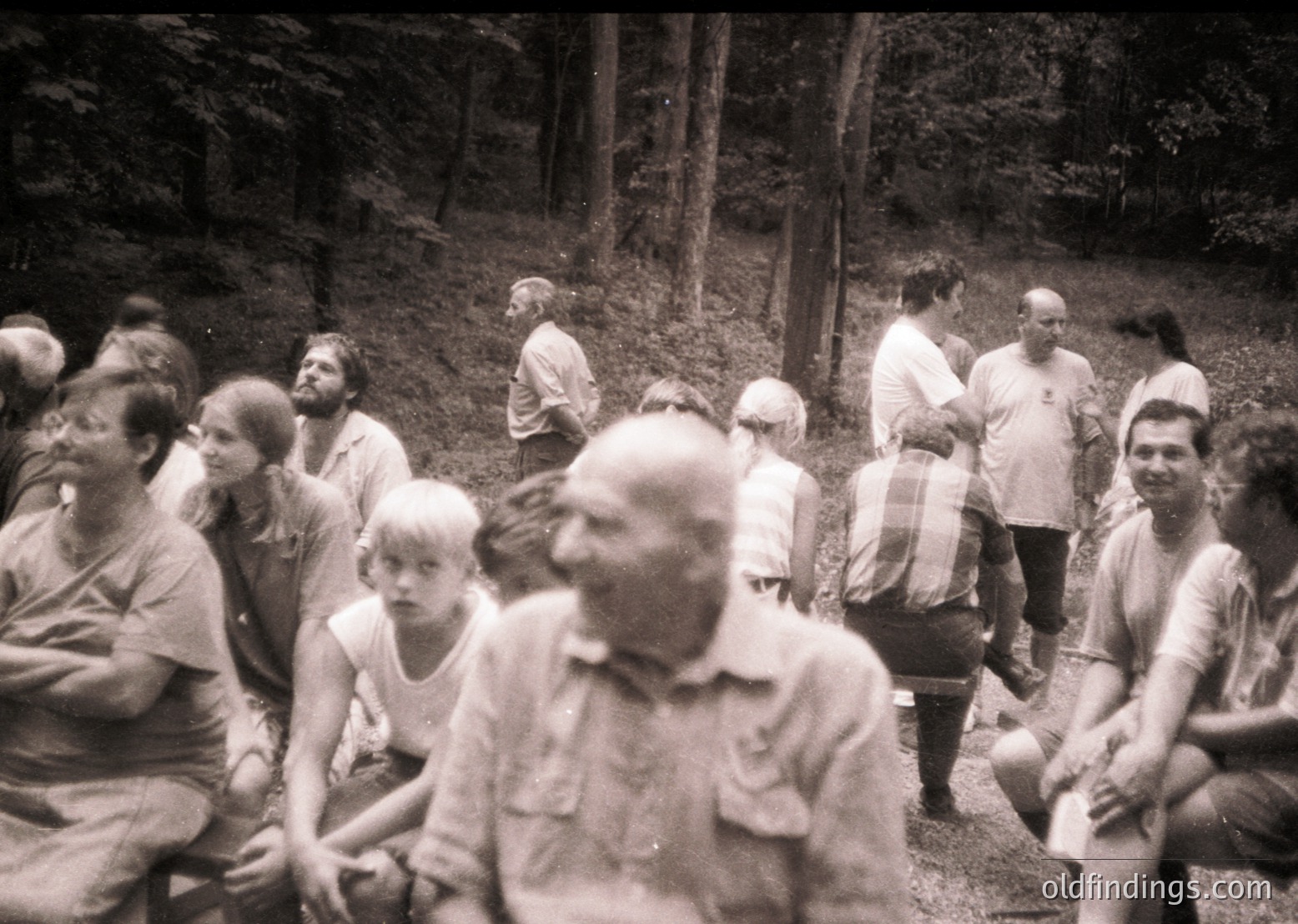 Group of people in a wooded area, likely mid-20th century. Casual summer attire suggests leisure or outdoor gathering. Dense forest backdrop with blurred motion indicates candid, informal setting.
