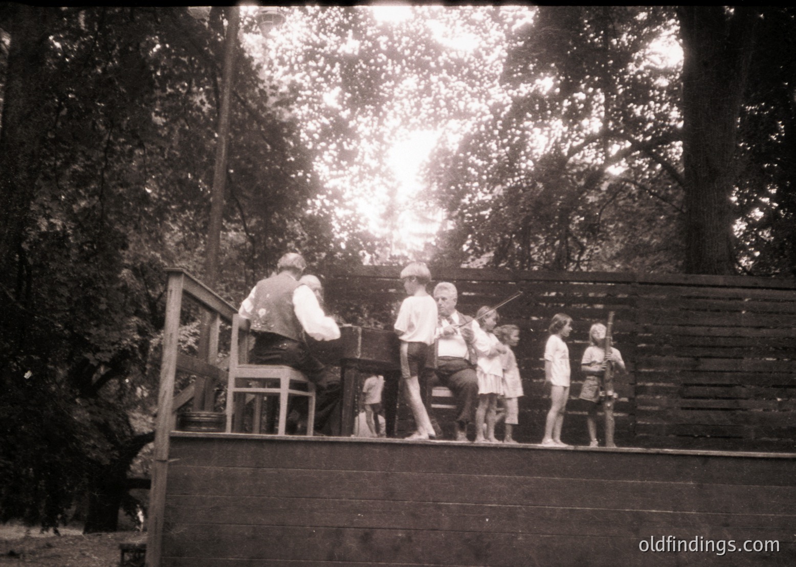 Vintage black-and-white photo of a group of children and an adult on a wooden deck surrounded by dense forest. The adult appears to be teaching or demonstrating piano playing. Mid-20th century (likely 1950s–1960s) outdoor music lesson setting.