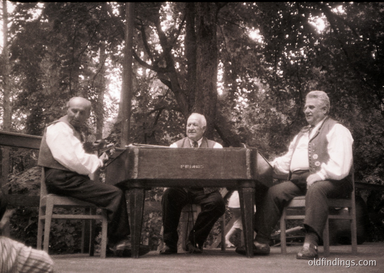 Three men in mid-20th-century attire perform outdoors near dense forest. Left: seated pianist playing a vintage upright piano. Center: conductor in suit directing. Right: male vocalist in vest with medal. Wooden stage and wooden benches suggest a rustic or park setting.