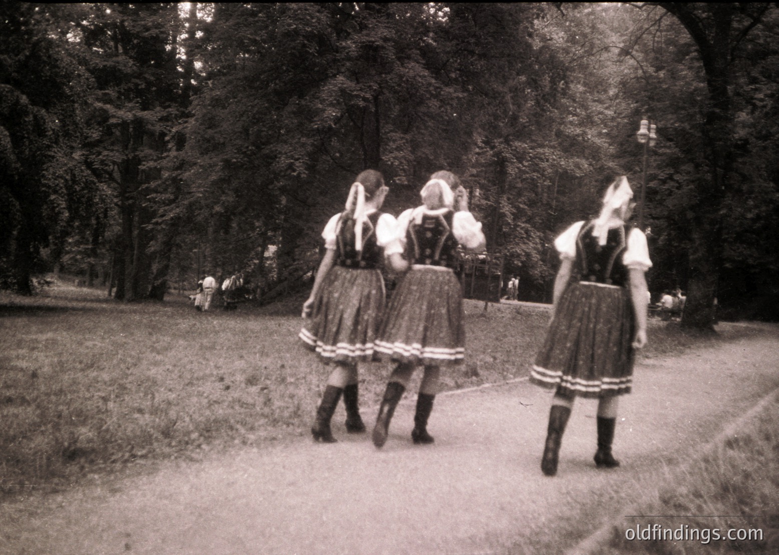 Three women in traditional Bavarian *Dirndl* dresses with white aprons and black knee-high socks walk through a park pathway, 1950s–1960s. Lush greenery and vintage sepia tone enhance nostalgic charm.