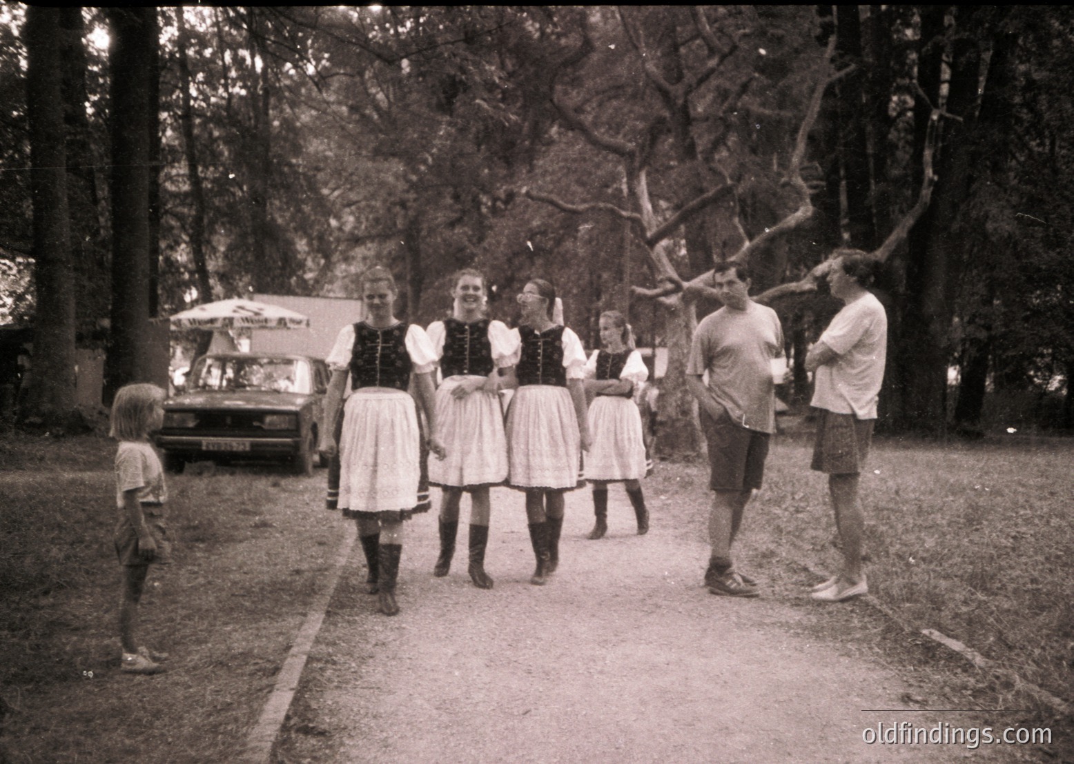 Mid-20th century outdoor gathering in rural setting. Five women in traditional folk attire (blouses, aprons, long skirts) and two men in casual shorts pose near a vintage van. Lush forest backdrop suggests a picnic or communal event, likely 1950s–1960s.