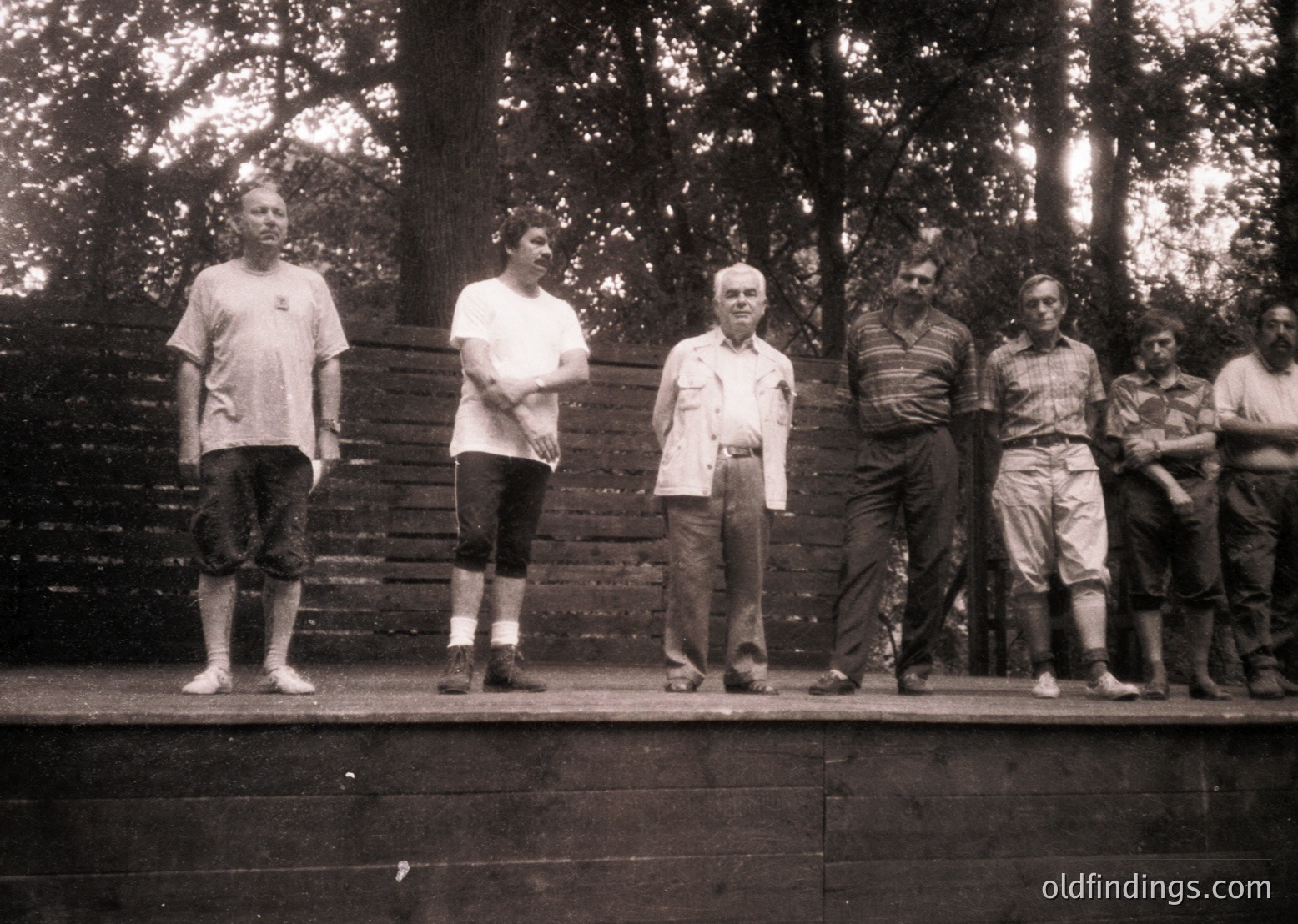 Black-and-white group portrait on a wooden platform, likely 1950s–1960s. Six men in casual attire—short-sleeve shirts, rolled pants, and sandals—pose outdoors near a stone wall and dense forest. Central figure wears a light-colored shirt with a pocket handkerchief.