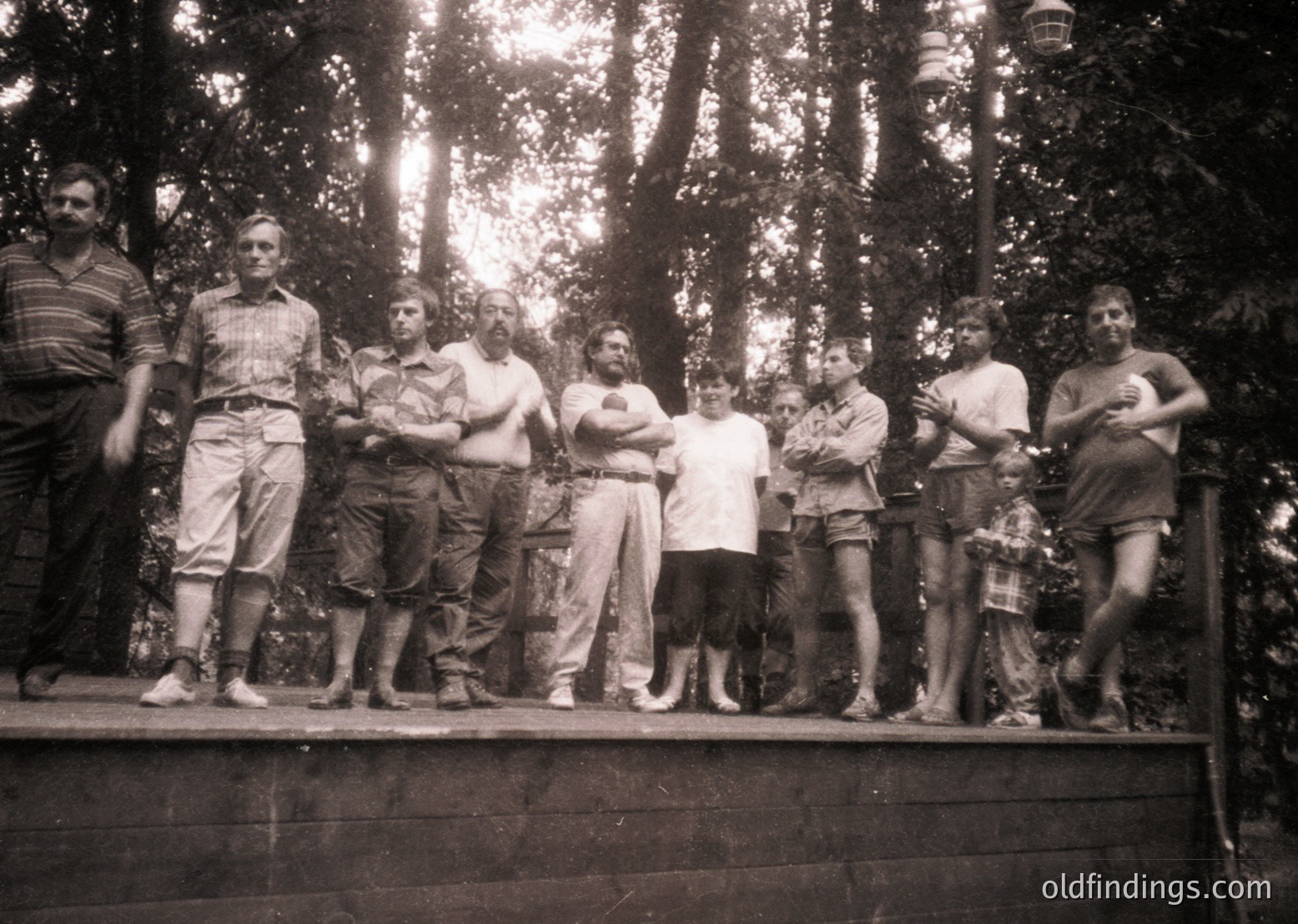 Black-and-white group portrait of eight men posing on a wooden platform in a forested area, likely 1960s–1970s. Casual summer attire includes short-sleeved shirts, shorts, and sandals. Dense pine trees frame the scene, with sunlight filtering through foliage.