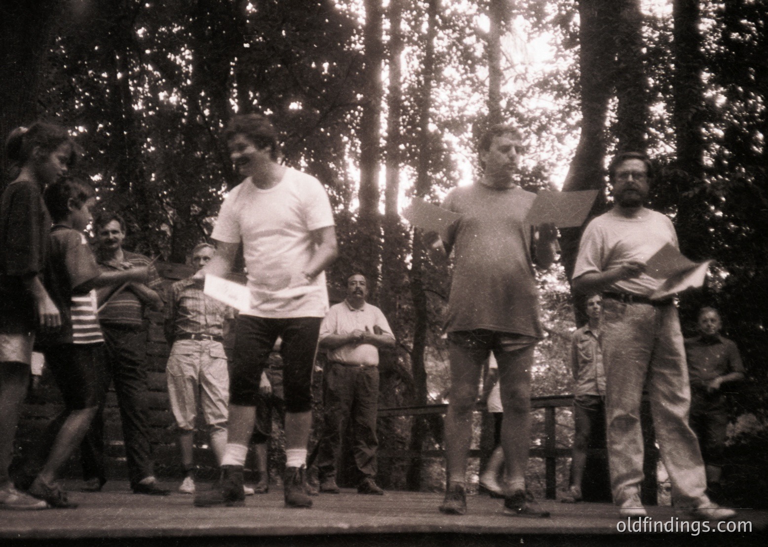 Black-and-white snapshot of a mid-20th-century outdoor performance. Three performers on a wooden platform, one in a white T-shirt and shorts mid-motion, another in a tank top with arms raised, and a third holding papers. Audience seated on benches behind them in a forested area. Casual 1960s–70s attire and grainy film texture suggest a spontaneous or amateur theatrical event.
