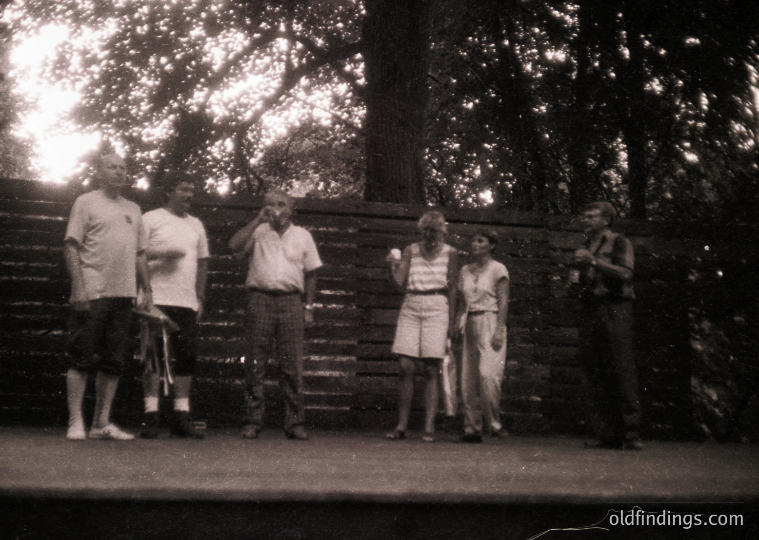 Vintage black-and-white group photo of six adults posing outdoors near a wooden fence. Men wear short-sleeve shirts and trousers; women in knee-length dresses. Mid-century fashion suggests 1950s–1960s. Natural light and blurred background emphasize candid, informal atmosphere.