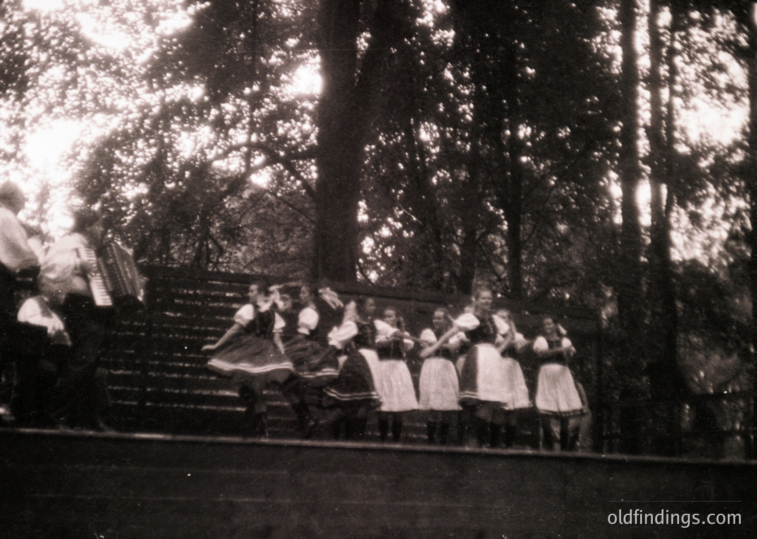 Vintage black-and-white photo of a folk dance ensemble performing outdoors in a wooded area. Women in traditional embroidered dresses and headscarves, arranged in a line on a wooden platform. Mid-20th century European folk culture, likely Eastern Bloc region.