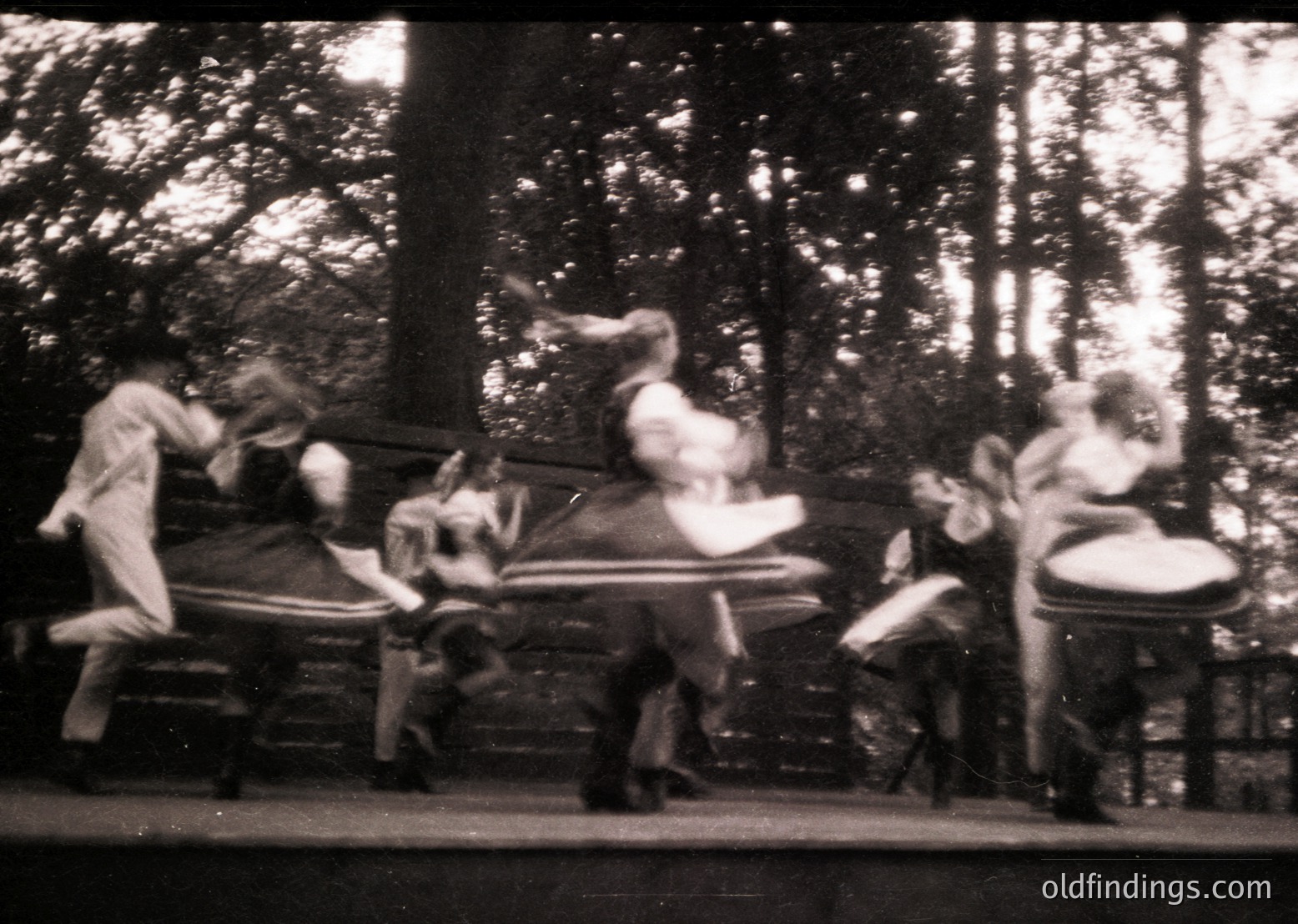 Vintage black-and-white photo of a carousel in motion, featuring blurred riders in 1920s-1930s attire. Wooden horses and a central carousel structure dominate the scene, surrounded by dense forest foliage. Ideal for historical or nostalgic stock imagery.