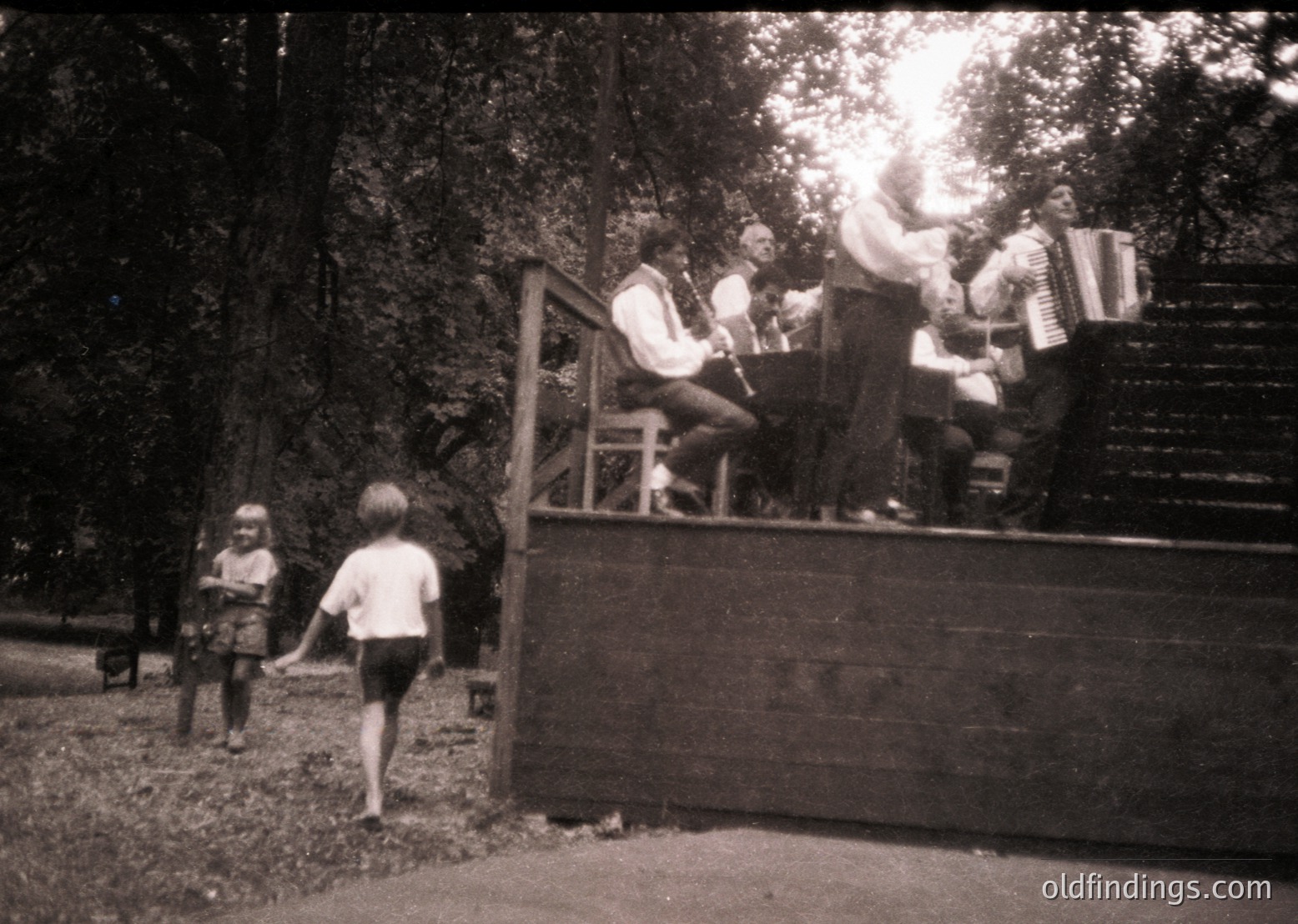 Vintage black-and-white photo of a folk band performing outdoors on a wooden platform, likely Eastern European . Accordionist, violinist, and vocalist engage the audience. A child in shorts walks toward the stage, while another child stands near a fence. Dense forest backdrop suggests a rural or park setting.