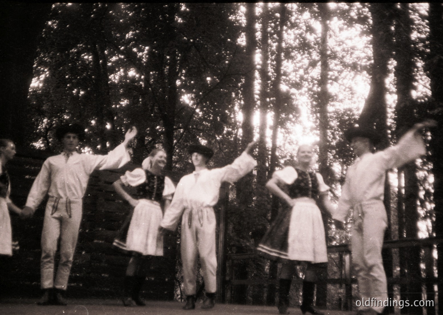 Group performing traditional folk dance in forest setting, likely Eastern European. Men in embroidered vests and women in long skirts with aprons, mid-20th century. Dynamic poses suggest lively cultural celebration.
