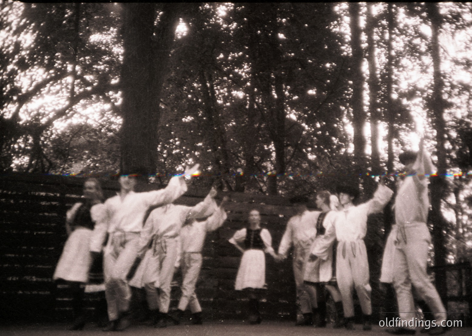 Vintage black-and-white photo of a traditional folk dance in a wooded area, likely Eastern European. Group of 7 dancers in matching white garments with dark belts, holding hands in a circle. Sunlight filters through leaves, creating dappled light patterns.