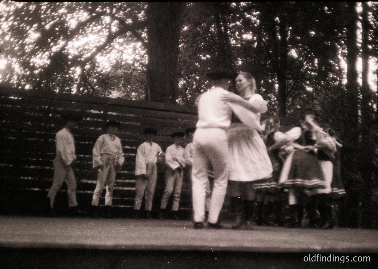 Vintage black-and-white photo of a folk dance performance in a wooded park setting, likely mid-20th century. Couple in traditional attire dances while others observe from wooden benches. Costumes suggest Eastern European cultural heritage.