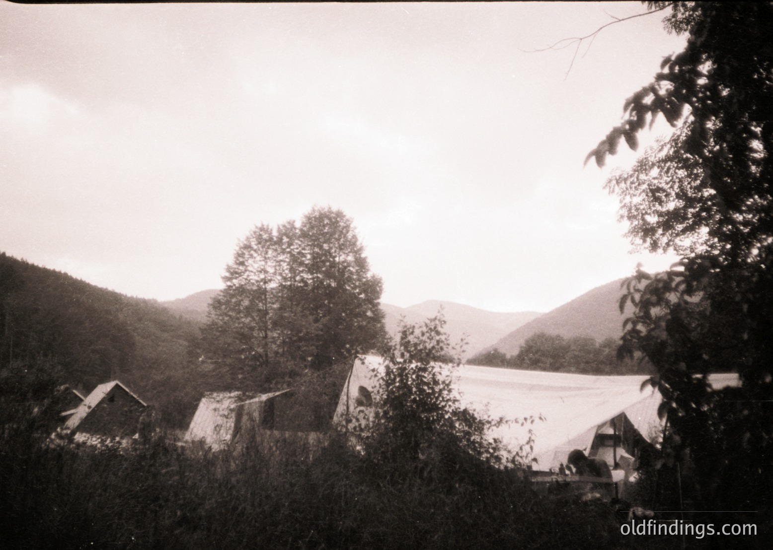 Vintage black-and-white photo of a rural campsite in mountainous terrain, featuring canvas tents and simple wooden structures. Dense foliage frames the scene, with misty hills in the background. Likely mid-20th century outdoor recreation or military use.