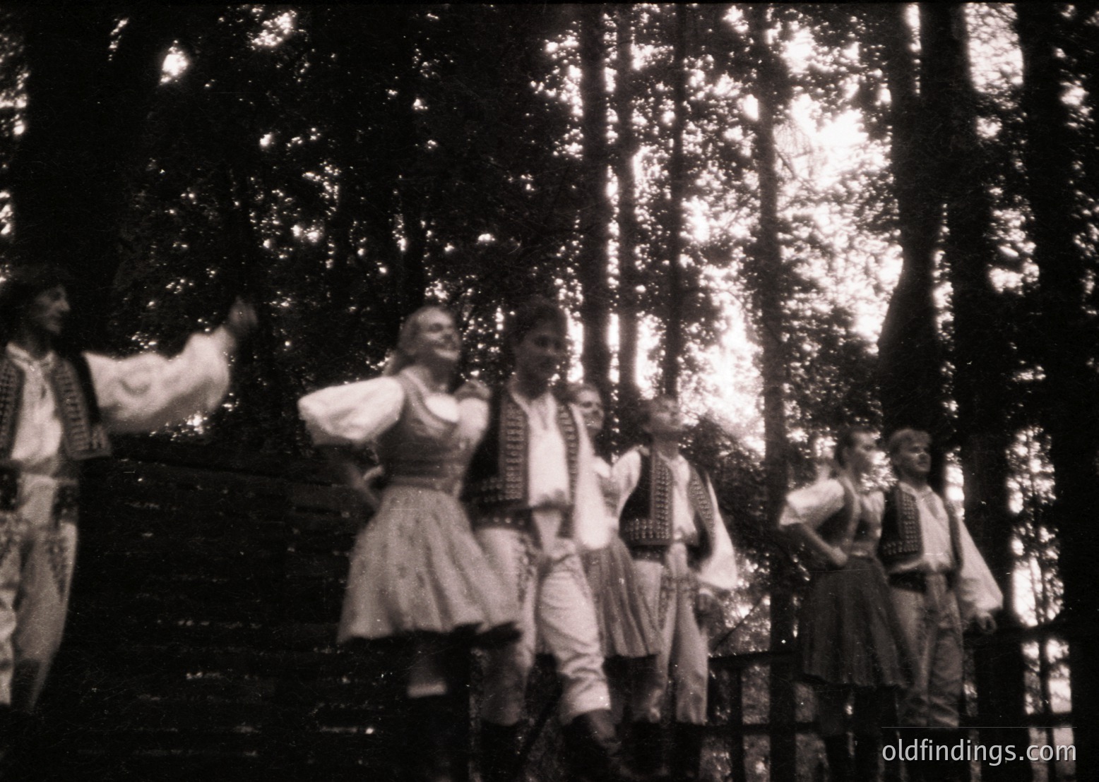Vintage black-and-white photo of a traditional folk dance performance in a forested setting. Six dancers in period costumes—men in embroidered vests, knee-length pants, and headbands; women in blouses, skirts, and headscarves—pose dynamically. Costumes suggest Eastern European heritage. Likely 1950s–1970s.